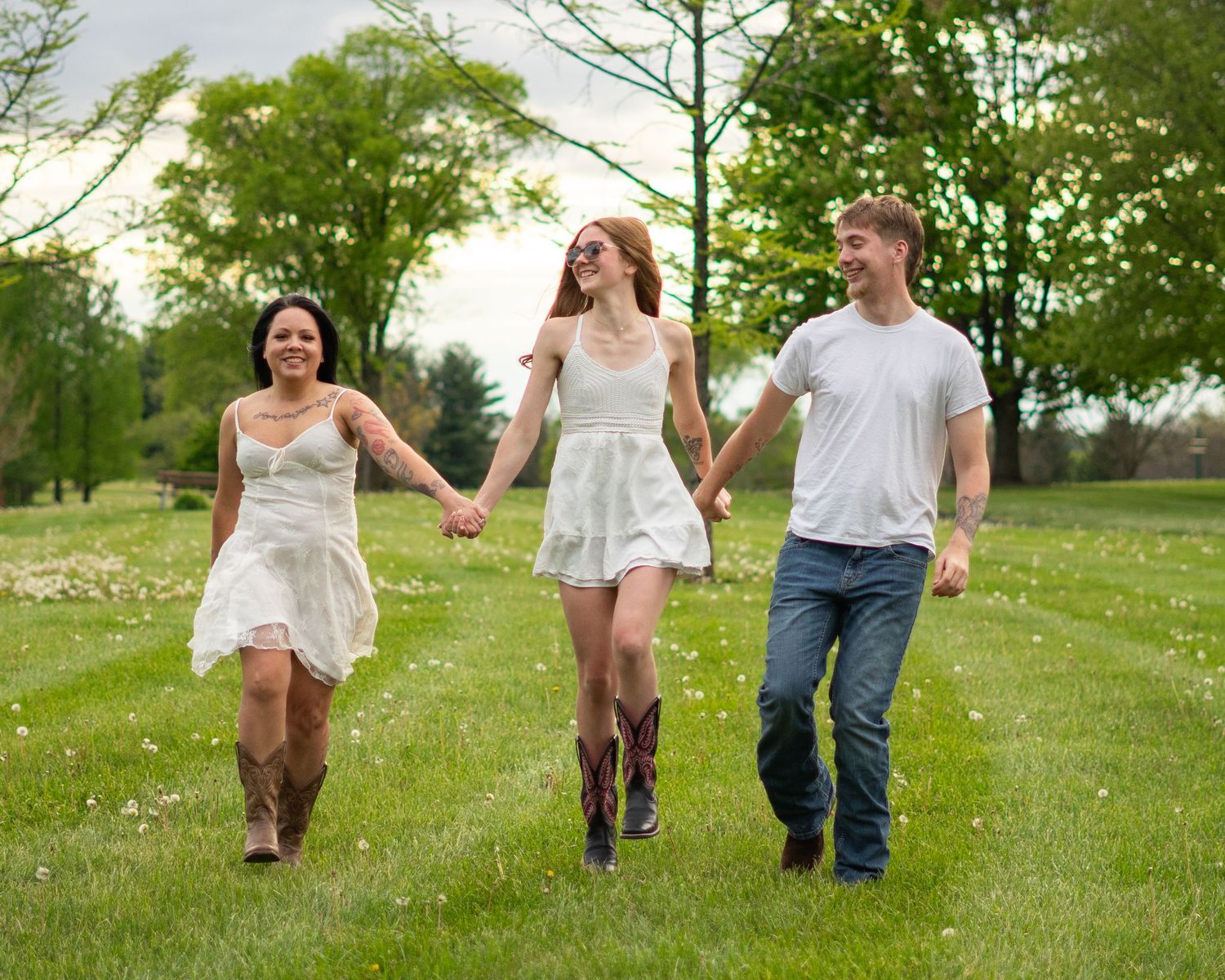 Three people holding hands walking through a lush green park, two women in white dresses and a man in jeans.
