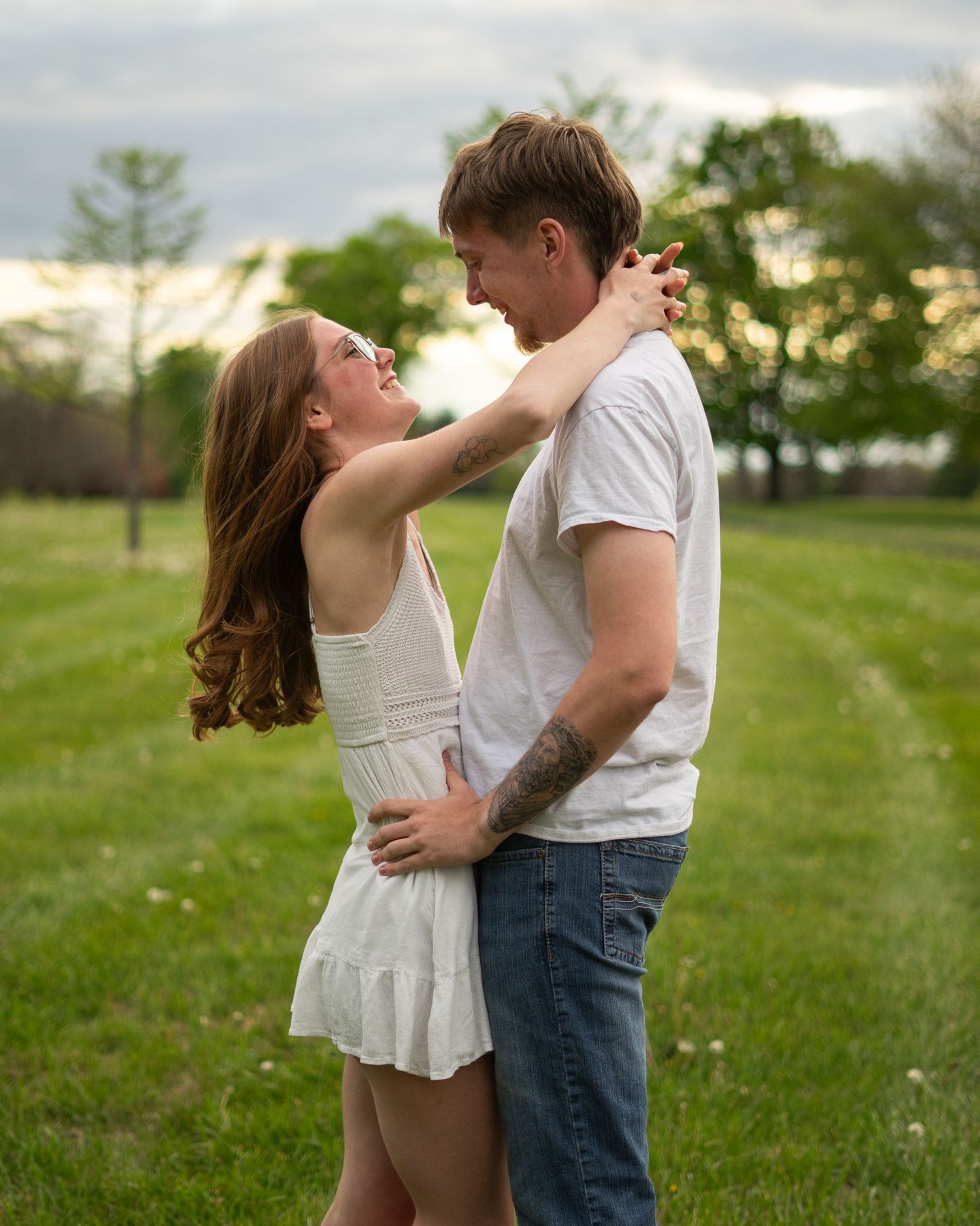 A couple in white outfits embraces romantically in a lush green field at sunset, the woman gazing up at the man.