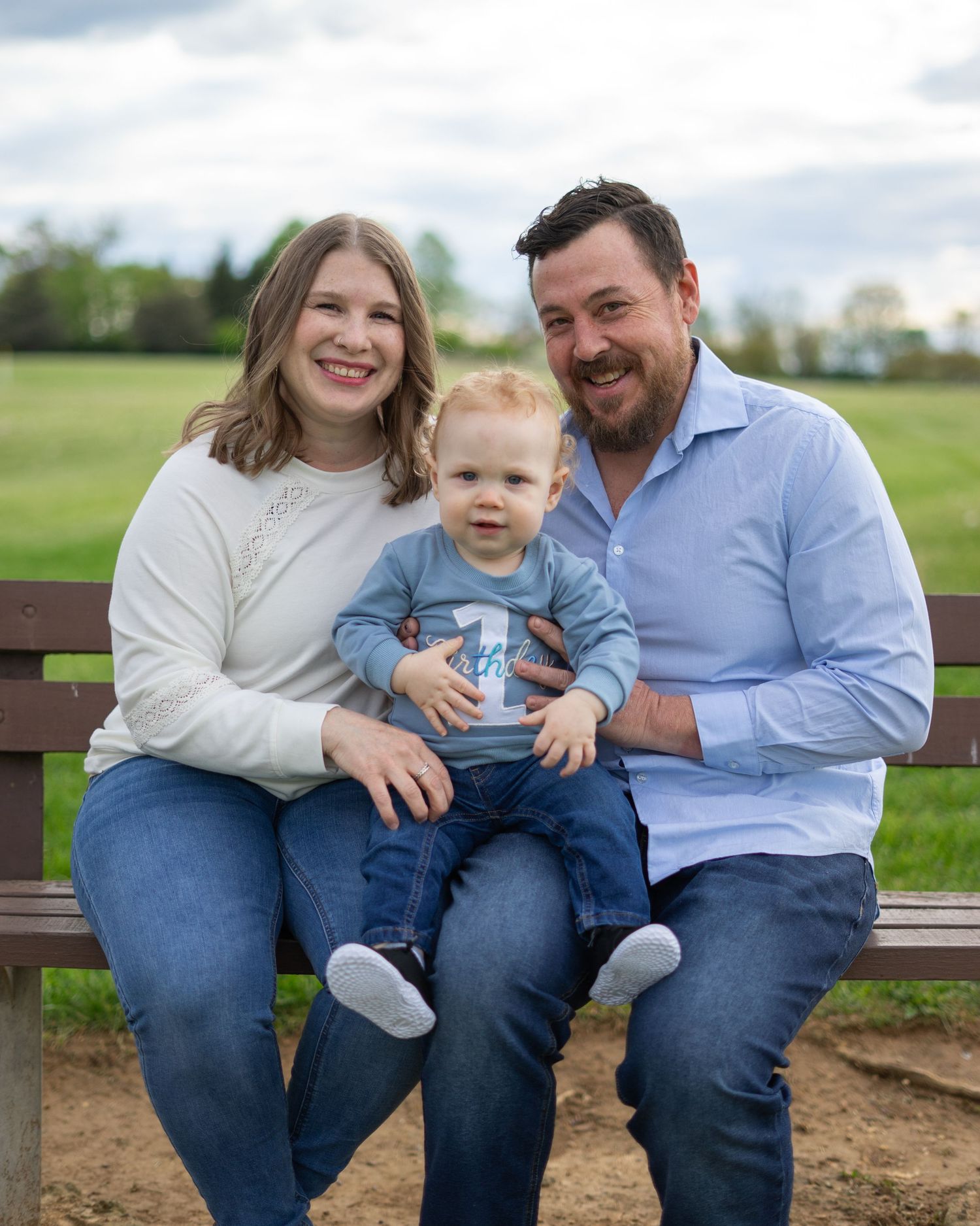A smiling family of three sits on a park bench outdoors, with parents flanking their baby dressed in a blue outfit.