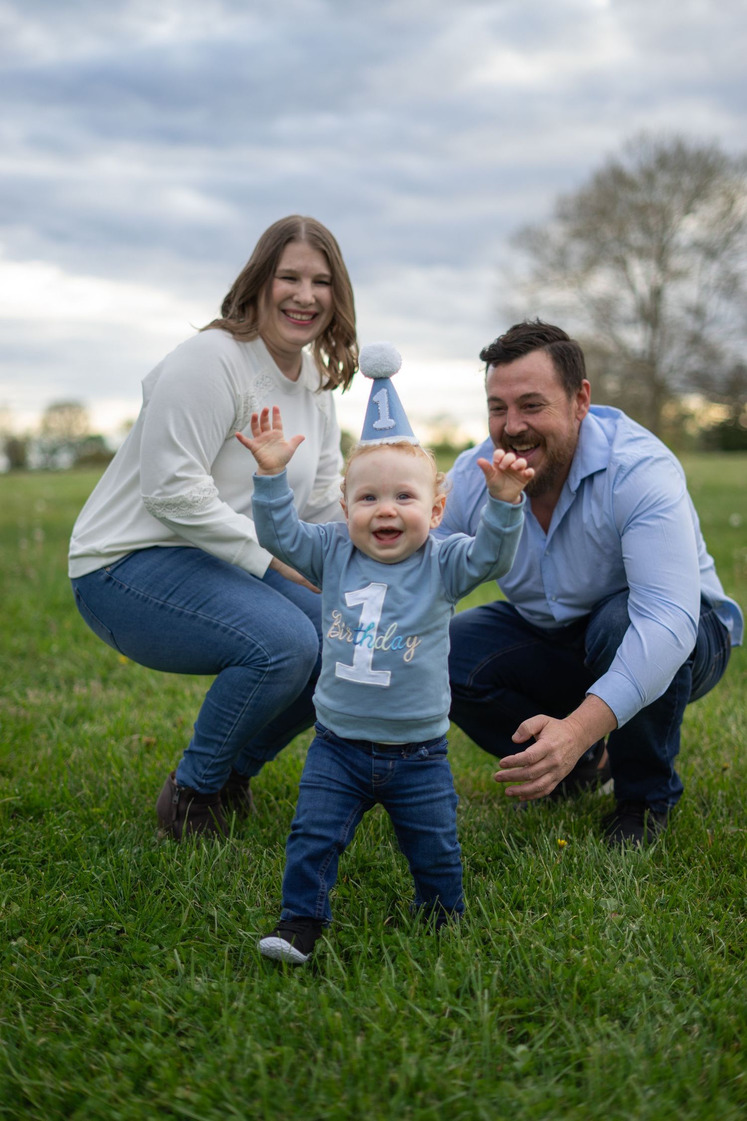 Family celebrating baby's first birthday outdoors on grass, parents crouching beside toddler wearing blue party hat.