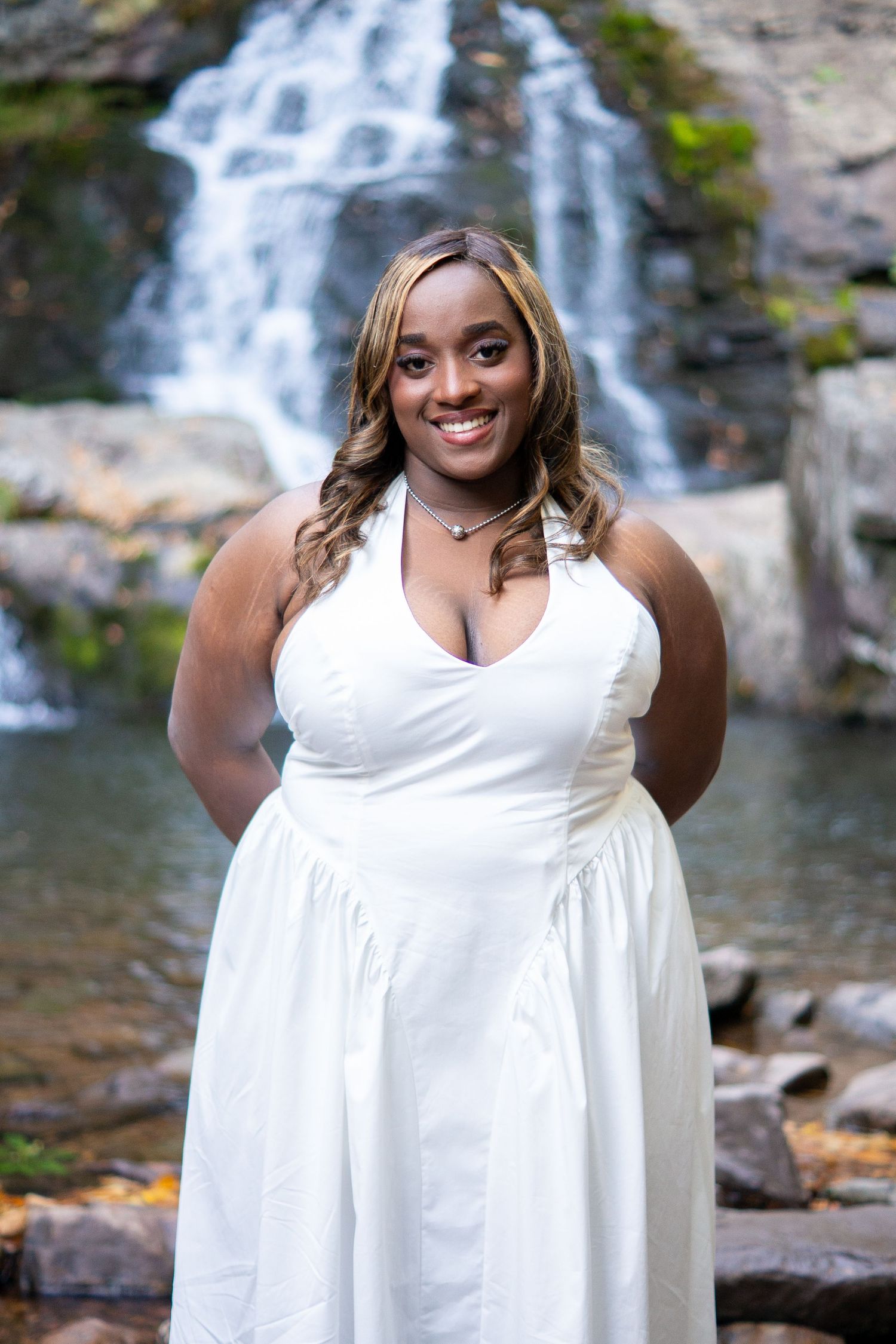 A person in a white dress poses in front of a cascading waterfall on a sunny day.