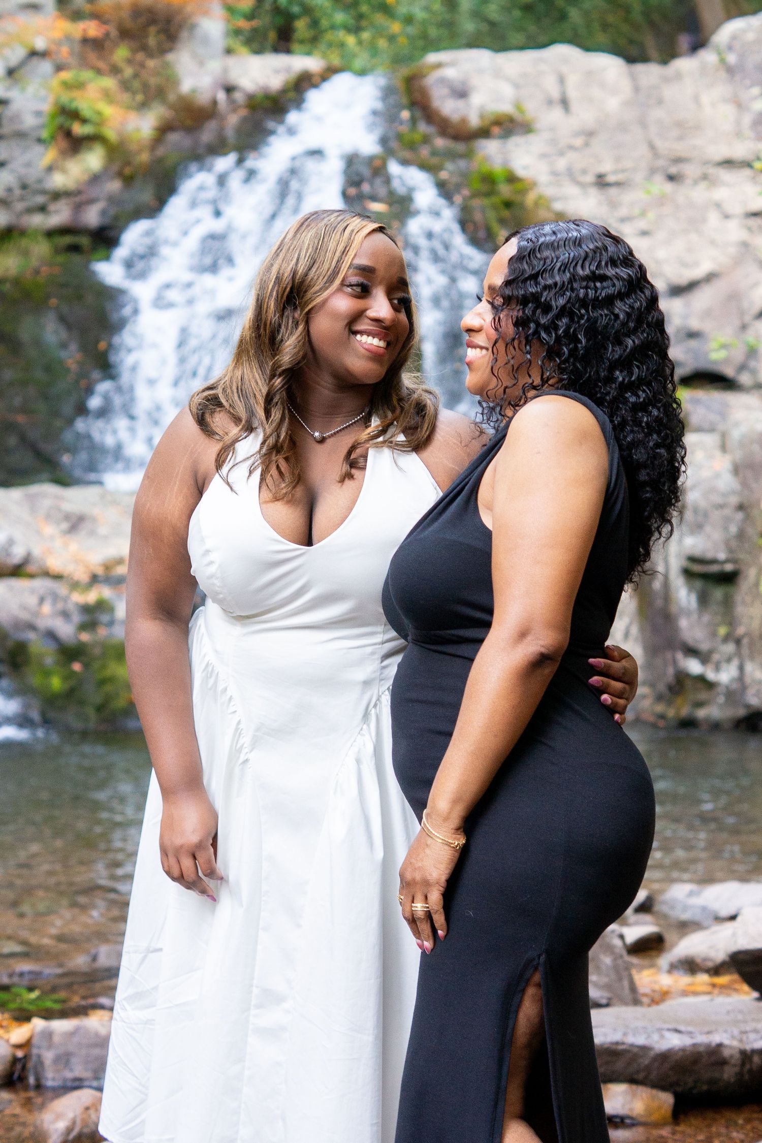 Two people in elegant dresses share a joyful moment by a waterfall.