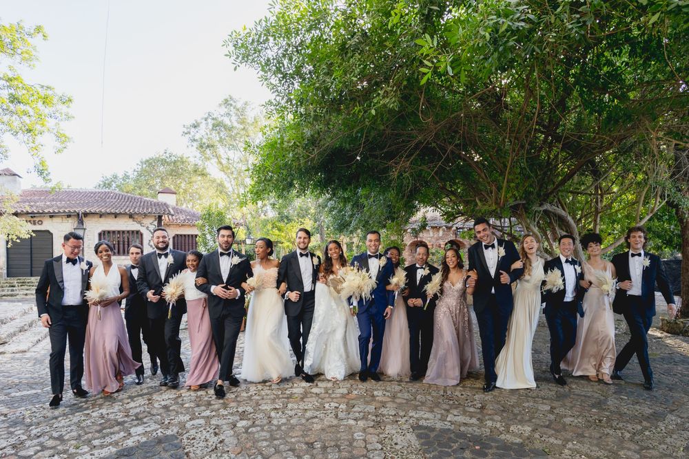 A large wedding party in formal attire poses together on a stone pathway surrounded by lush greenery.