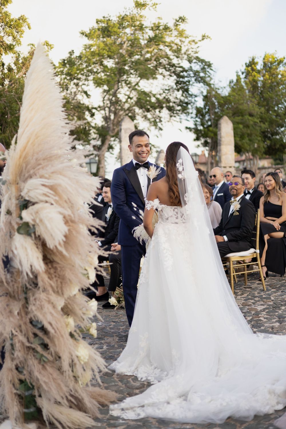 Elegant outdoor wedding ceremony with pampas grass decor and guests seated on wooden chairs.