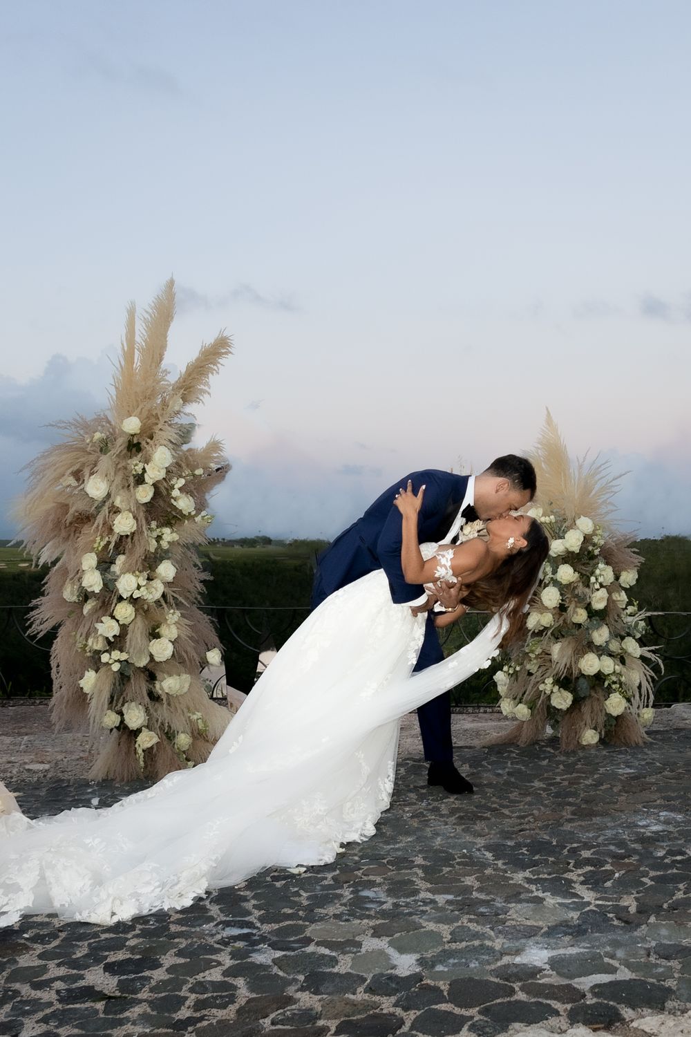 Romantic wedding couple poses in a dip kiss against a scenic backdrop with pampas grass and floral arrangements.