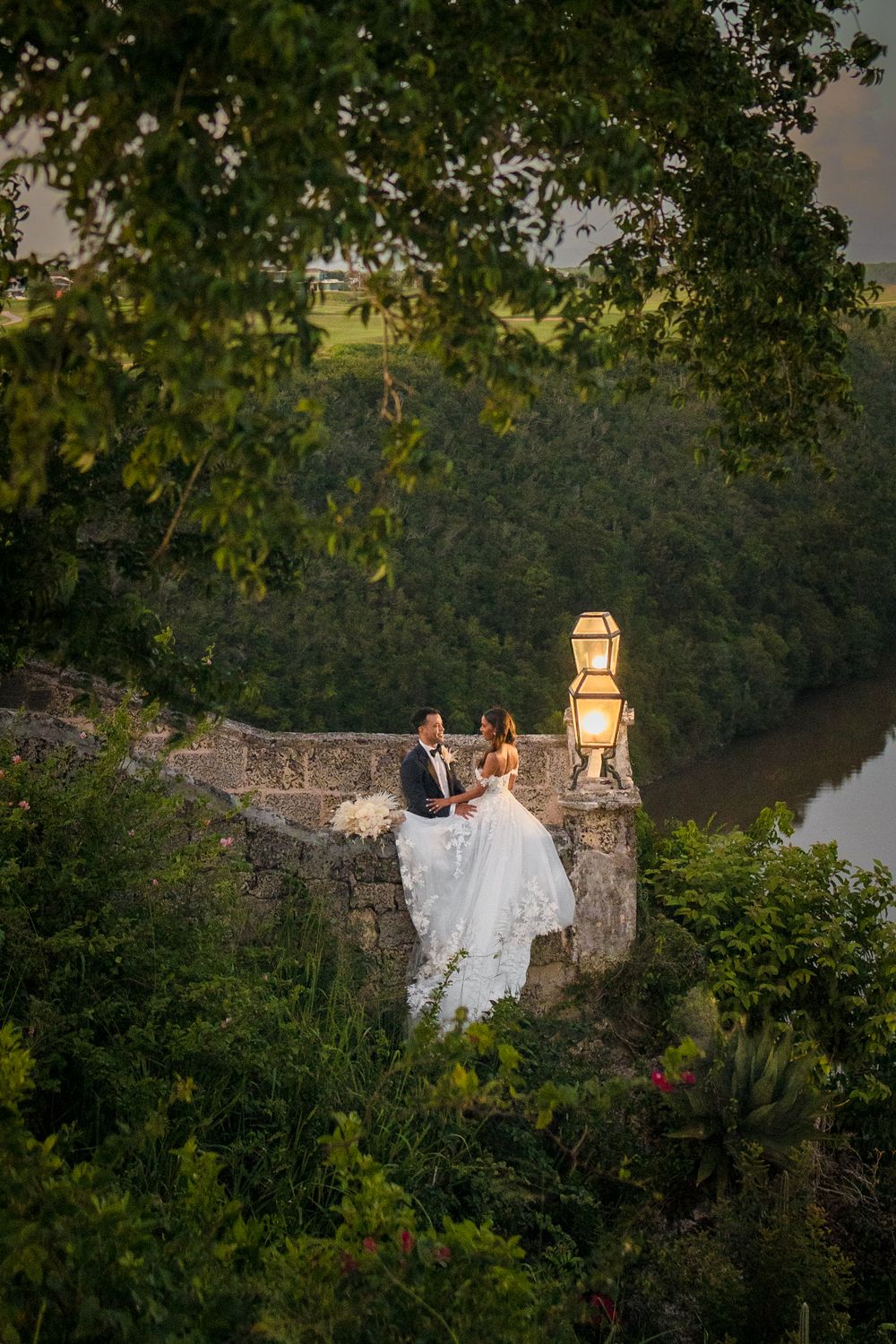 A couple in wedding attire sits on a stone wall overlooking a tranquil lake at sunset with lanterns glowing nearby.