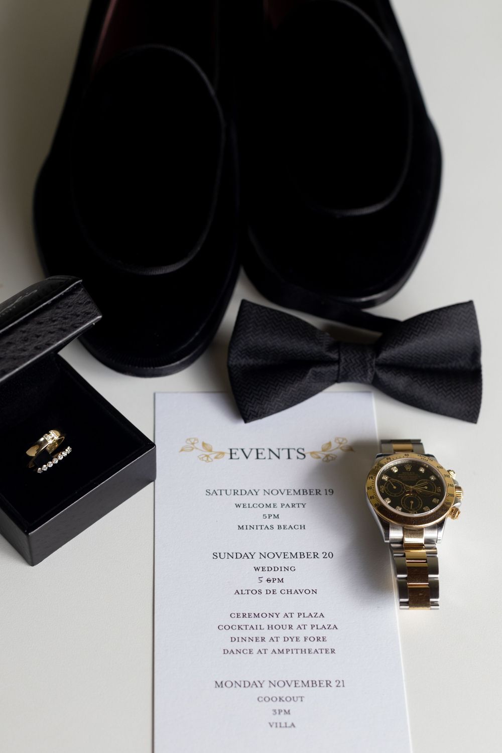 Wedding accessories including black bow ties, a gold watch, and invitation cards arranged on a white surface.