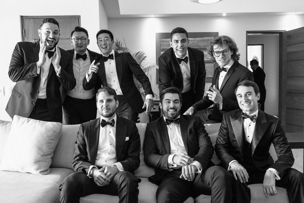Groomsmen in black tuxedos pose together for a formal wedding party photo in black and white.