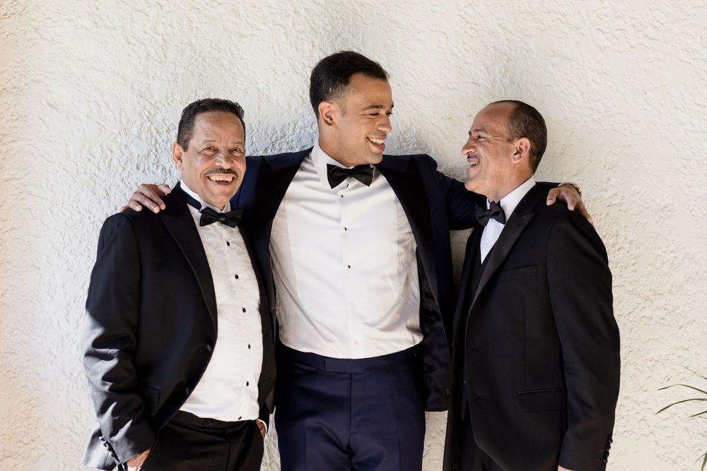 Three people in formal black and white tuxedos share a lighthearted moment together against a light colored wall.