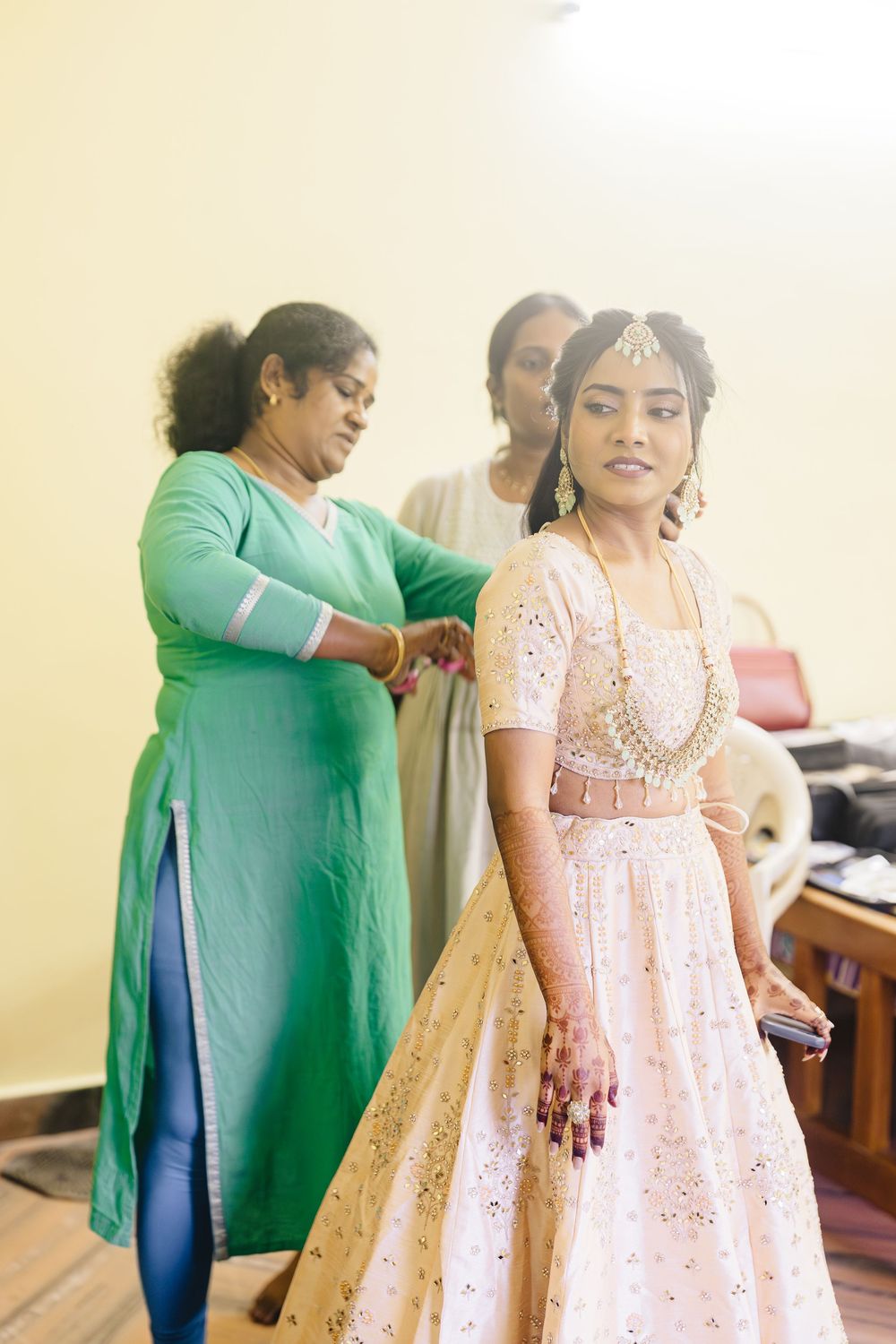 A woman in a green traditional dress helps another in a pink and gold lehenga prepare for a special occasion.