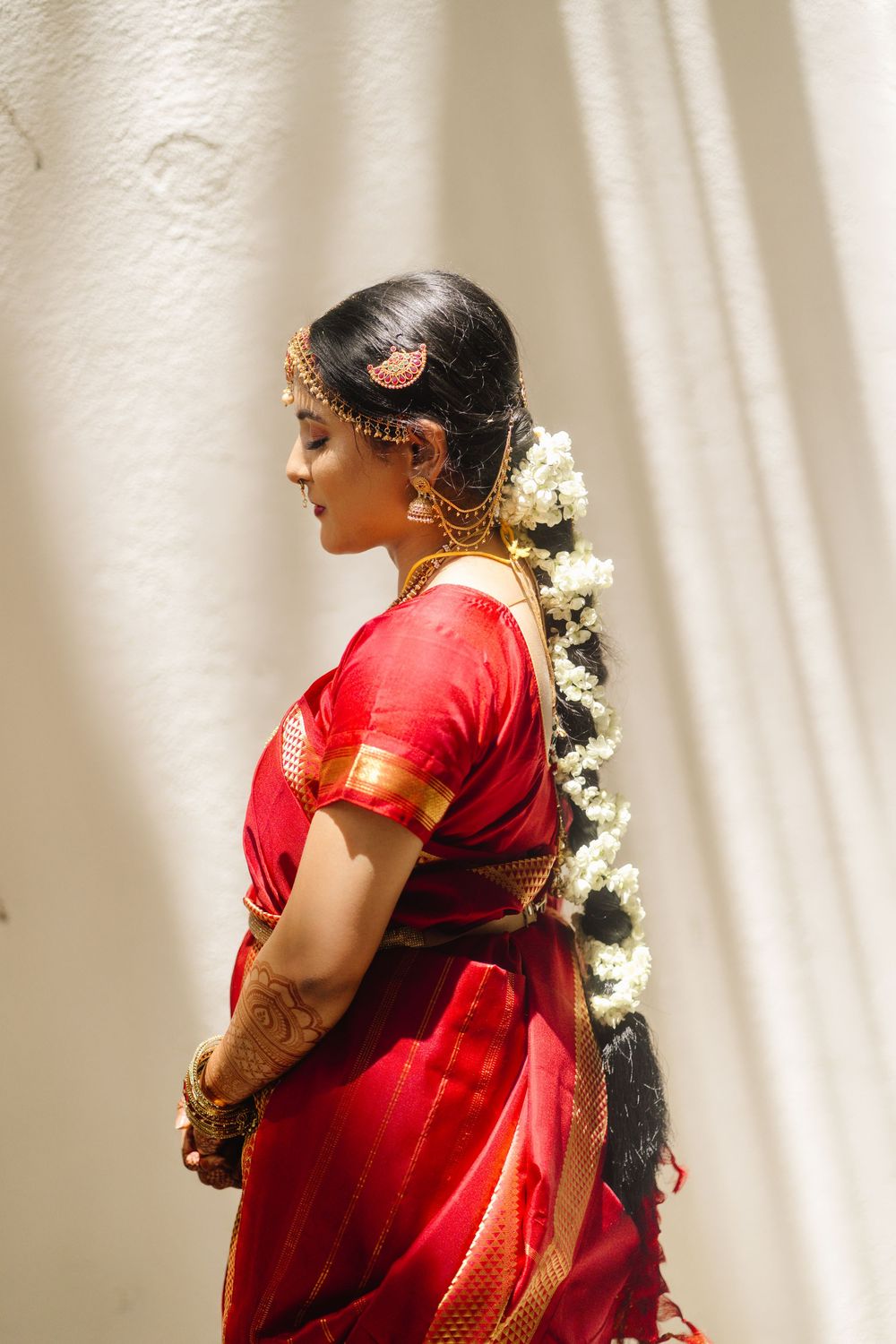 Side profile of bride in red silk saree with traditional gold jewelry and jasmine flowers in hair.
