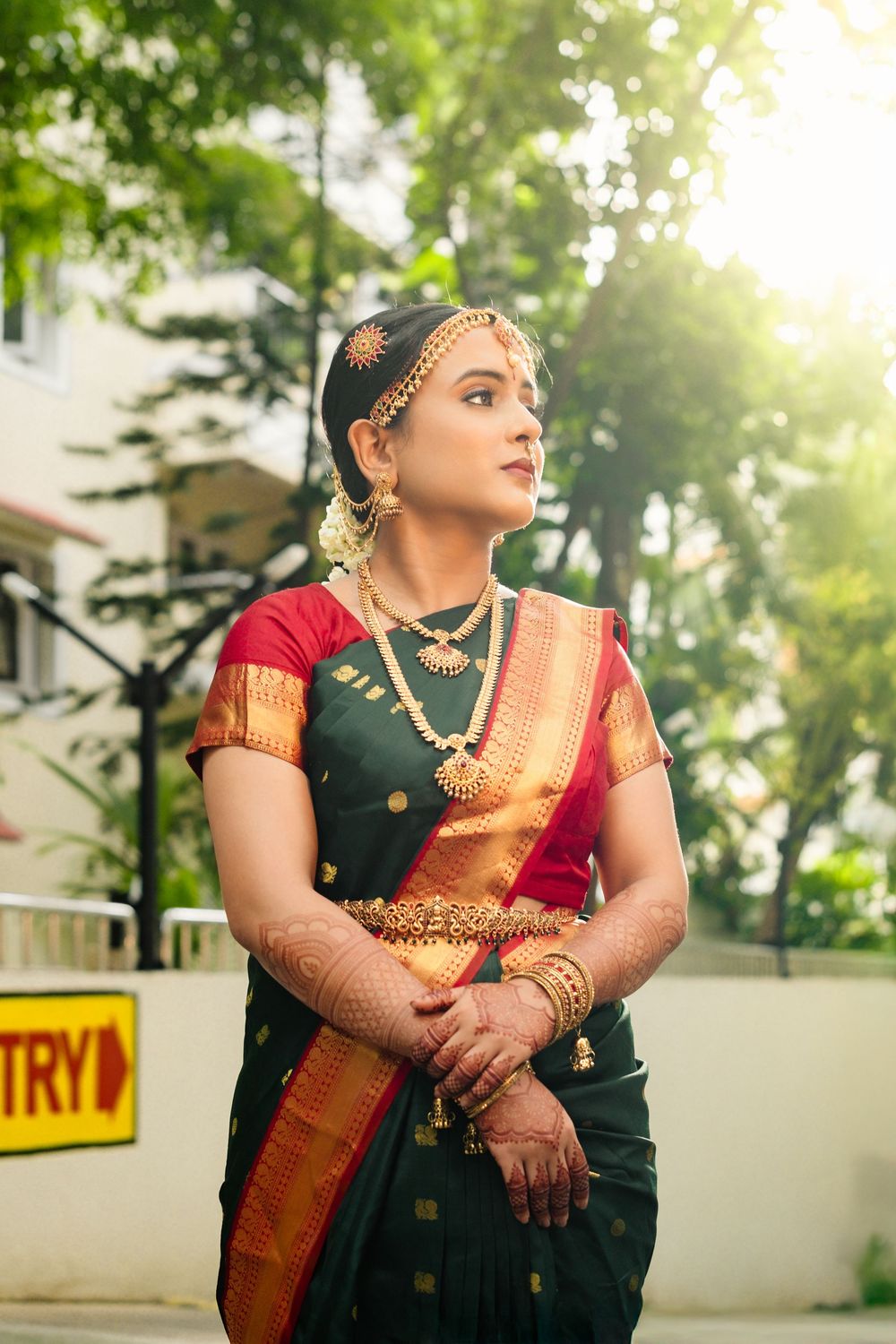 Traditional Indian bride in a dark green and red saree poses elegantly against a sunlit outdoor backdrop.
