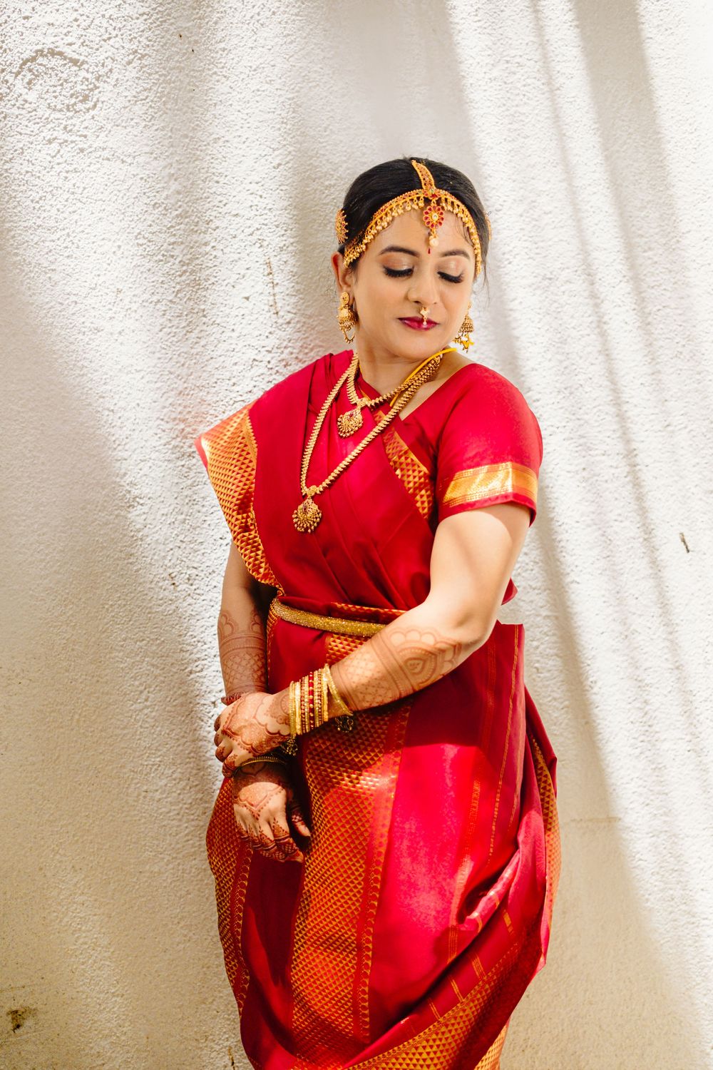 Traditional Indian bride in red silk saree with gold jewelry poses gracefully against white wall.