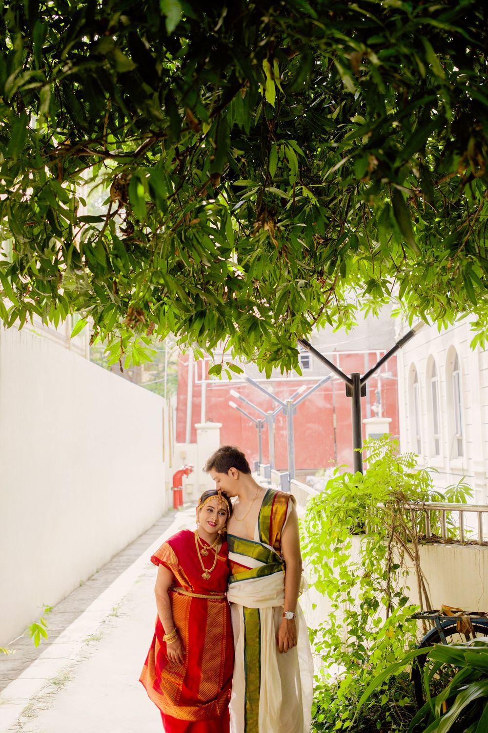 Couple in traditional red attire pose together under lush green foliage during outdoor photoshoot.