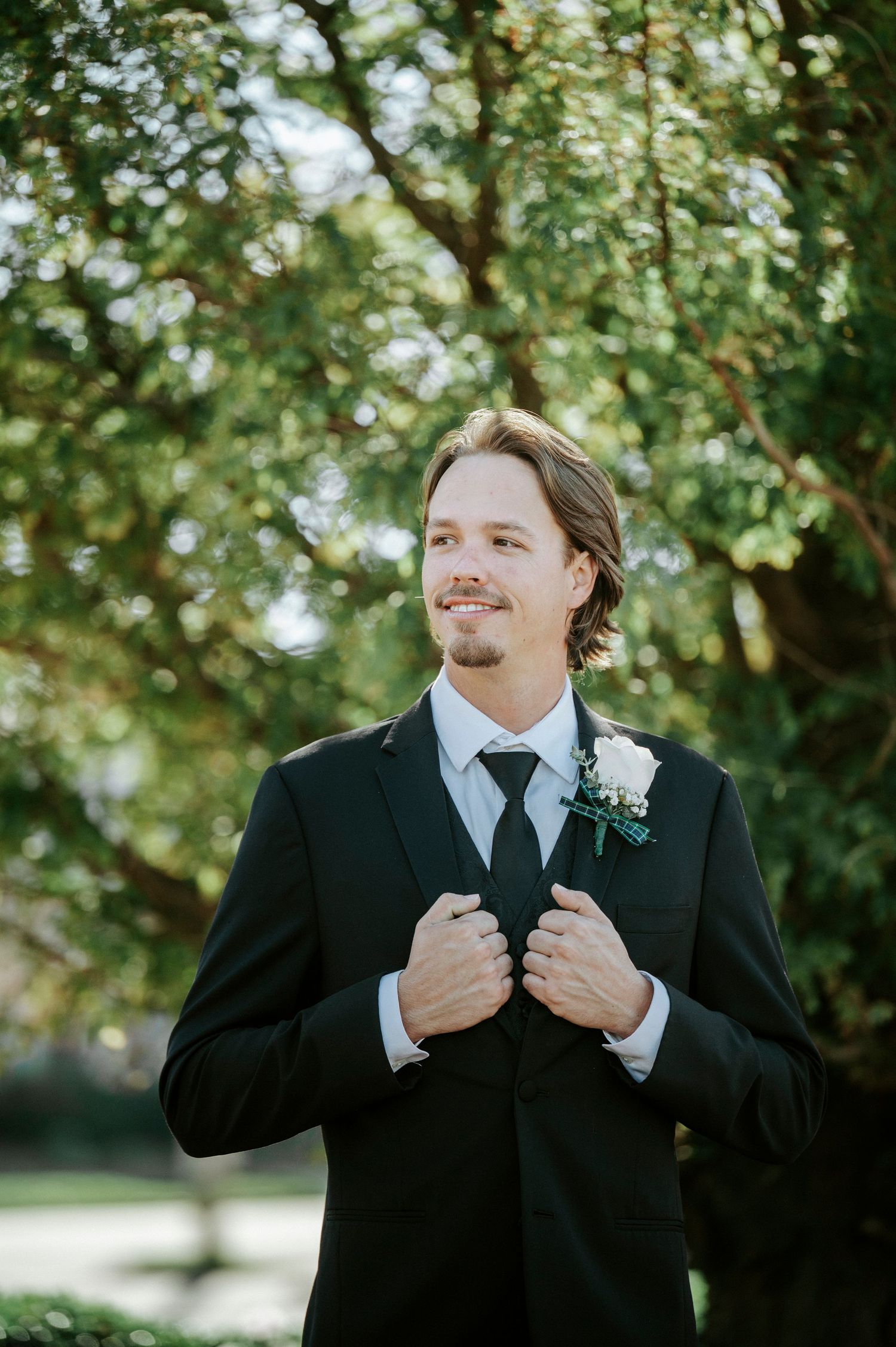A person in a black suit adjusts their tie while standing outdoors among trees in a natural setting.