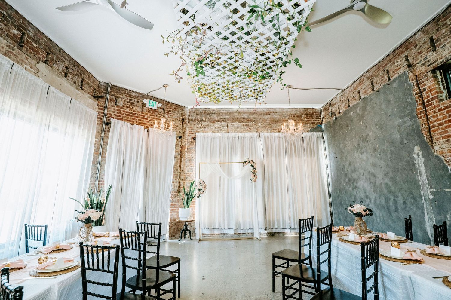 A rustic indoor wedding venue with exposed brick walls, white curtains, and hanging greenery decorations.