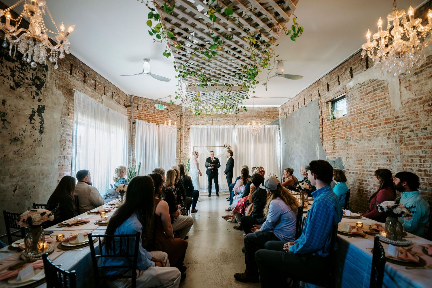 An intimate indoor wedding ceremony in a rustic brick room with hanging chandeliers and greenery on the ceiling.