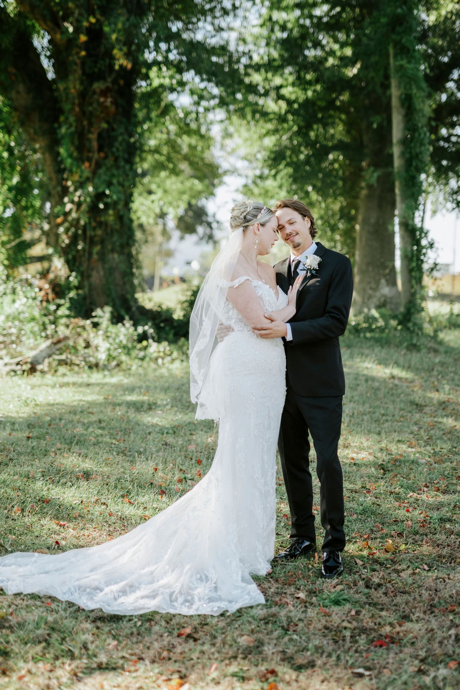 A couple in wedding attire shares an intimate moment on a grassy hillside under tall trees.