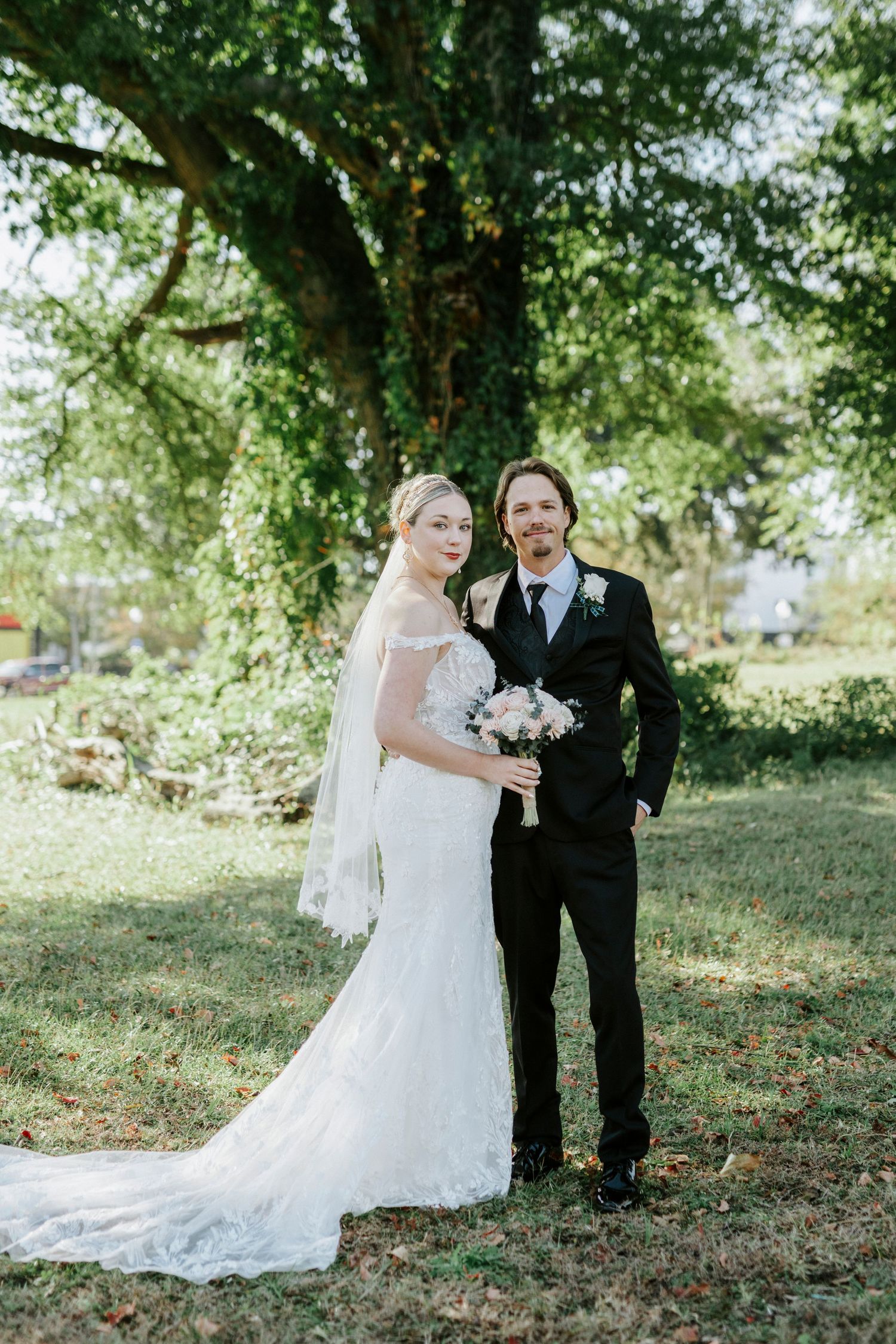 A couple poses for their wedding portrait outdoors under a large tree with lush green foliage in the background.
