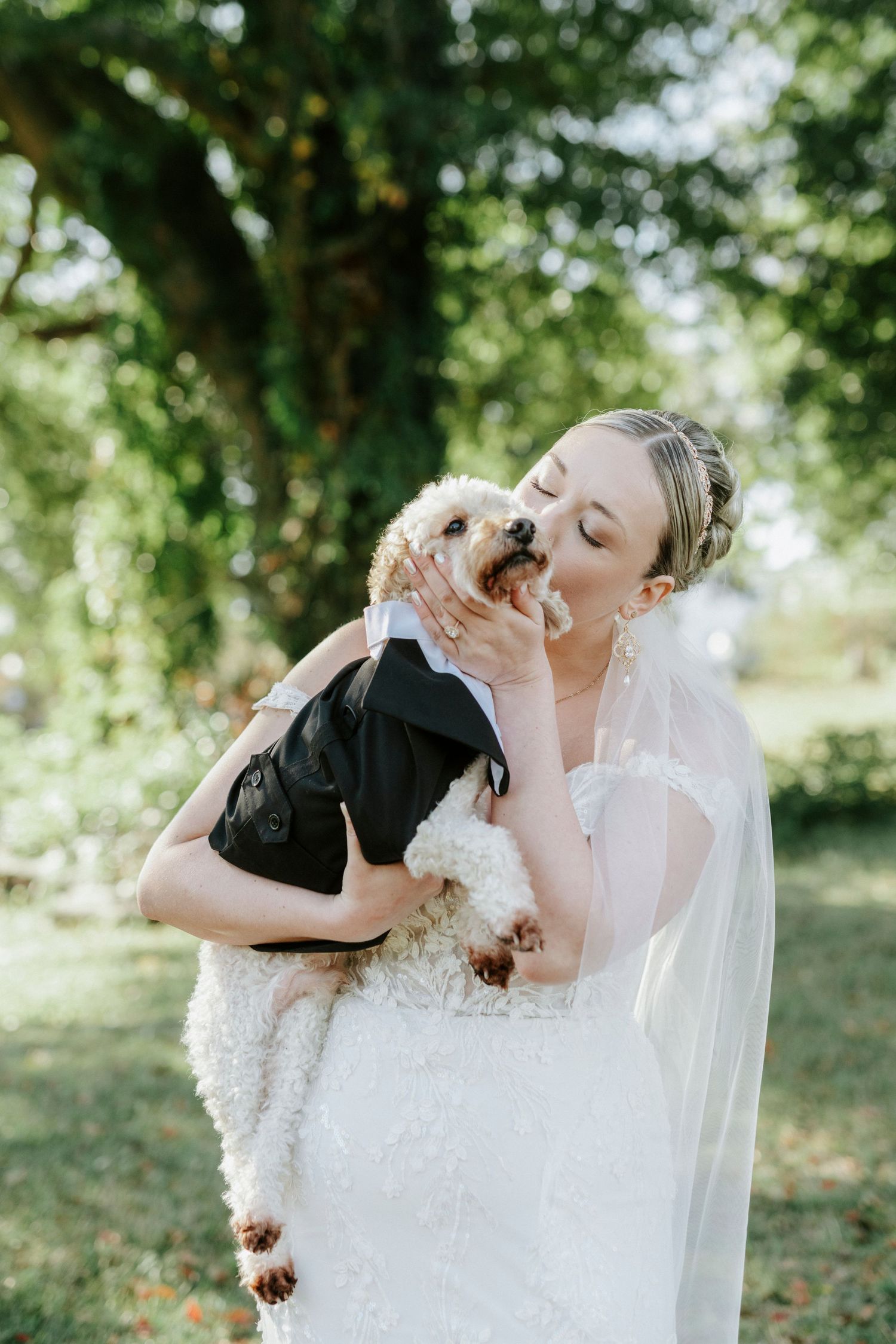 A bride in a white lace wedding dress lovingly embraces and kisses a black dog in a garden setting.