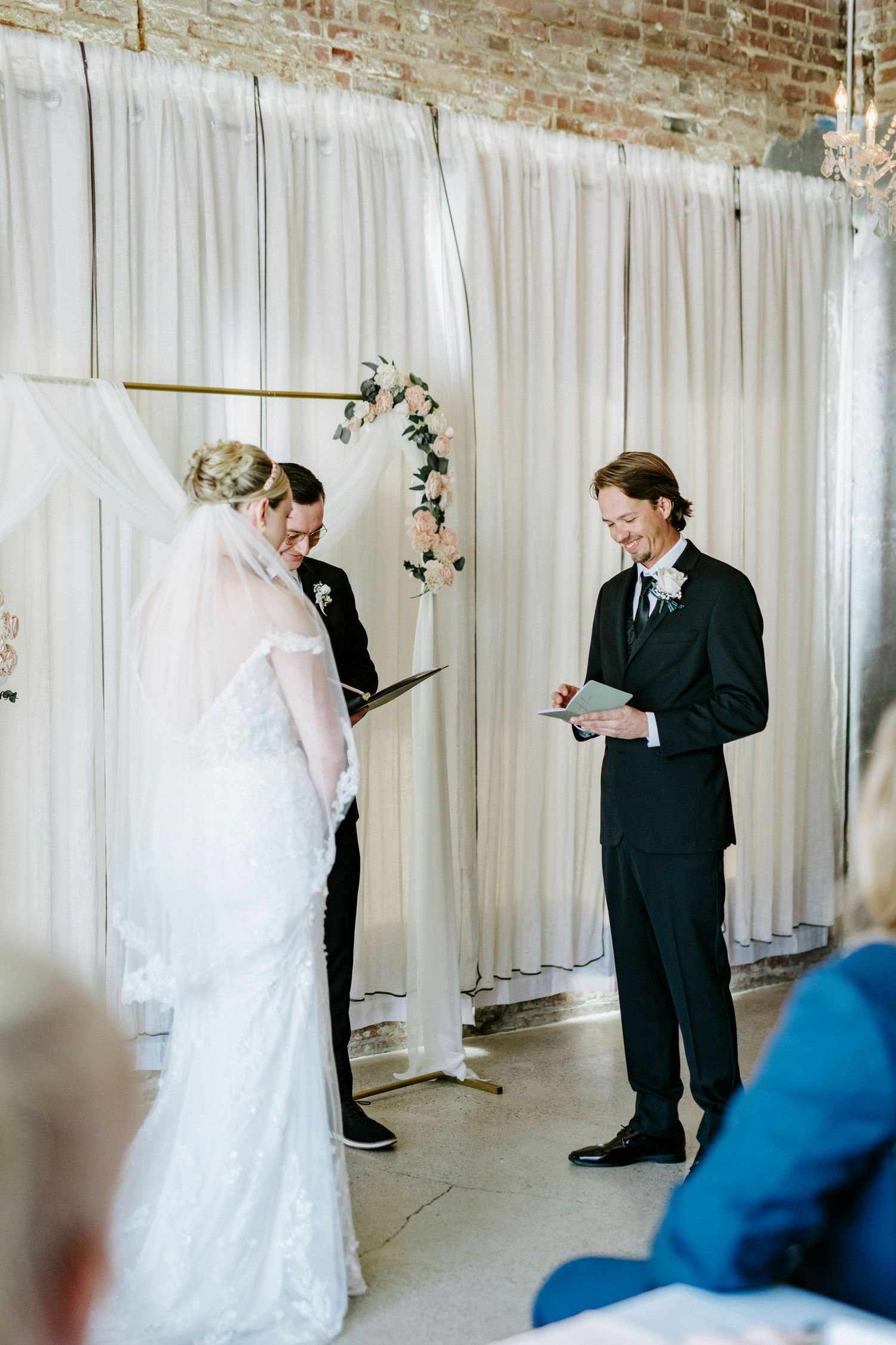 A wedding ceremony takes place in an indoor venue with exposed brick walls and white curtain backdrop.