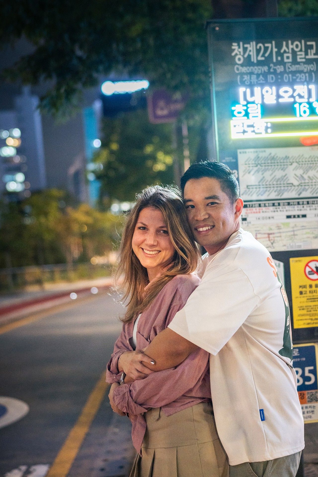 A couple shares a romantic moment at night in front of neon signs and Korean text on a city street.