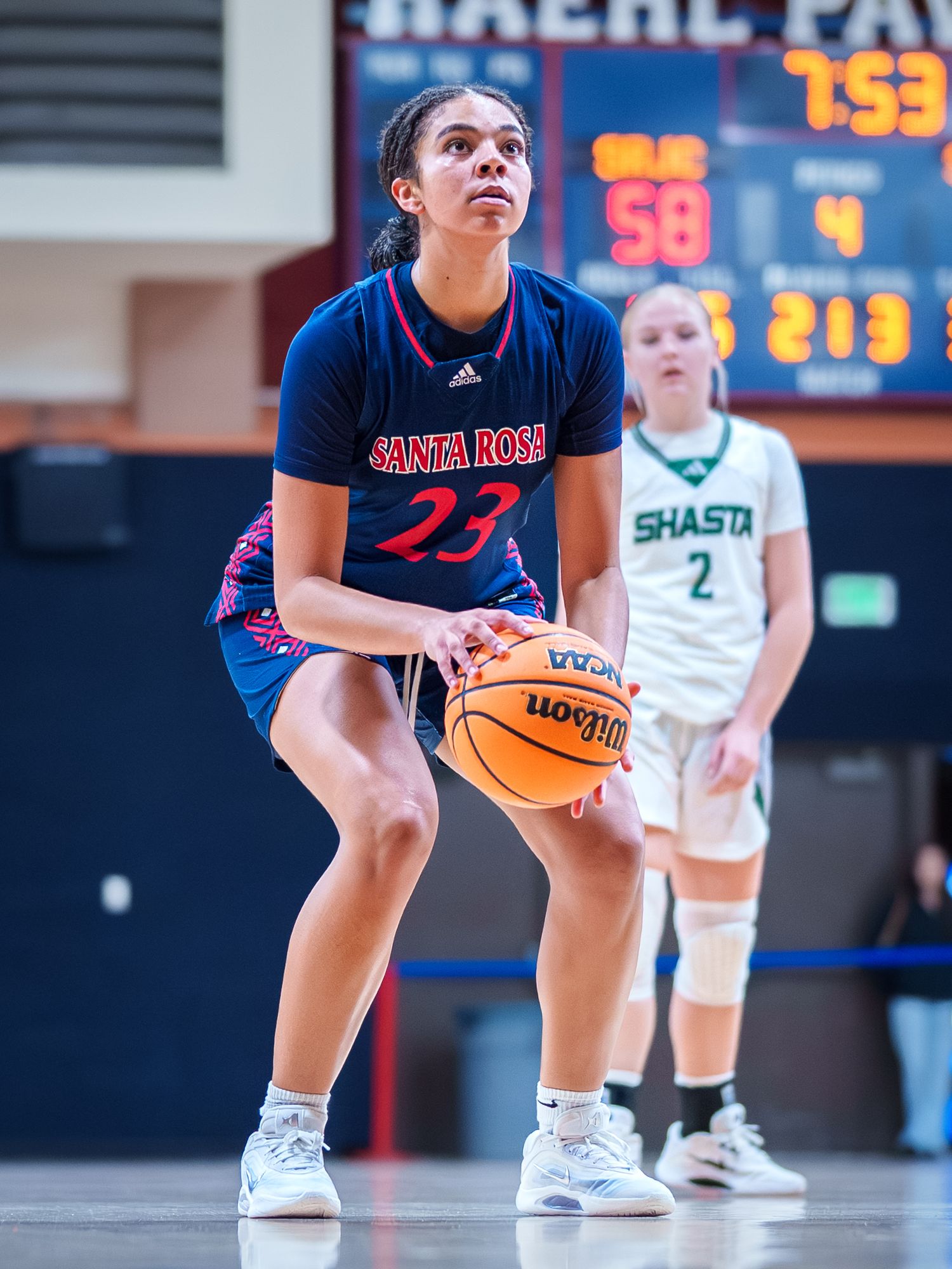Basketball player in navy blue uniform crouches low in defensive stance during game action.