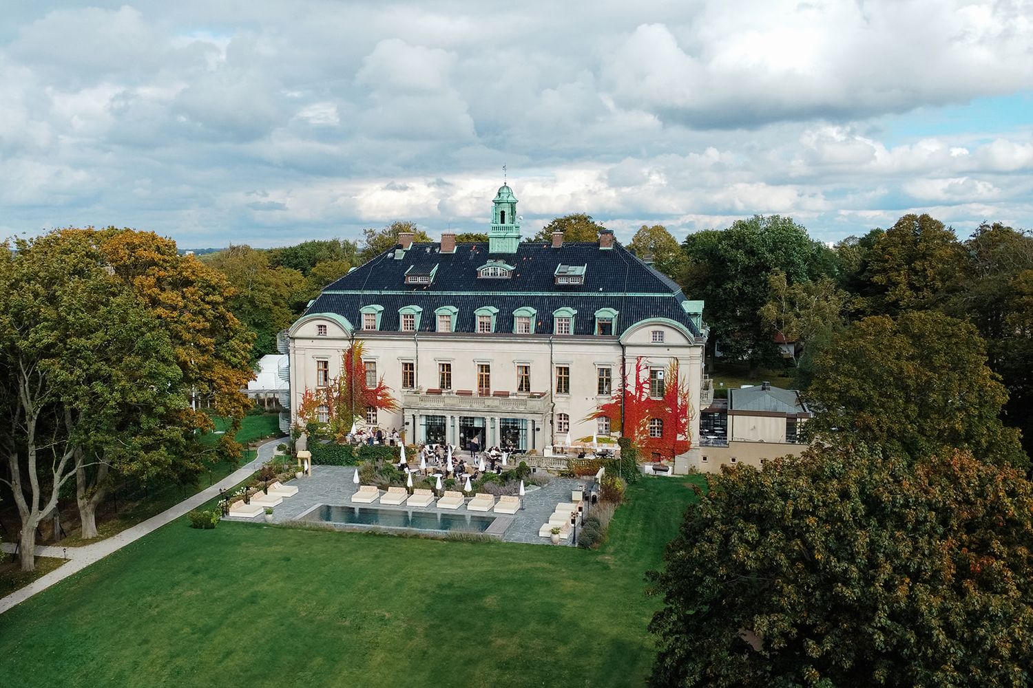 Panoramic view of a grand historic estate with white buildings and green landscaping surrounded by autumn trees.