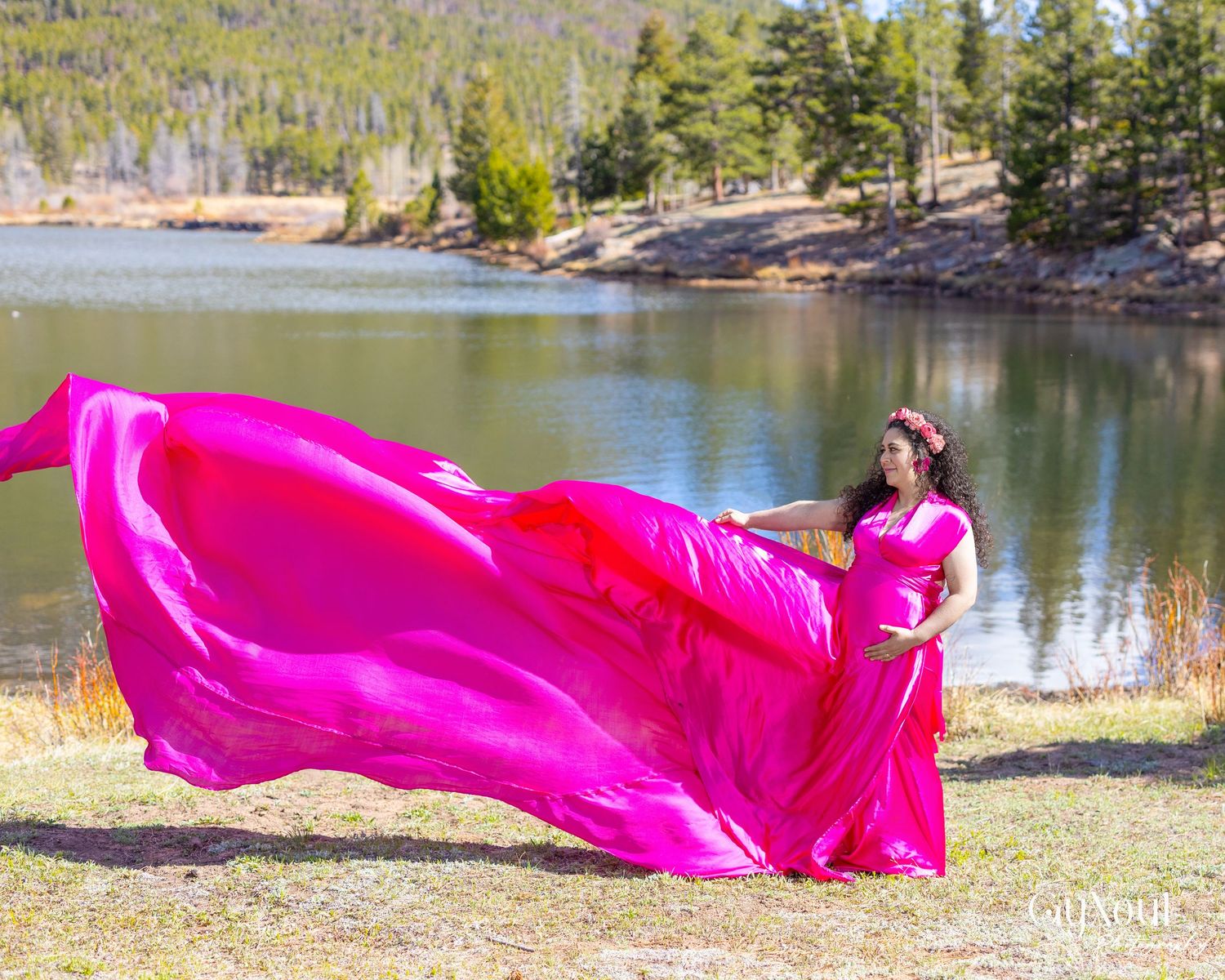 A bright pink silky fabric flows gracefully across a scenic lakeside landscape with pine trees and rocky shores.