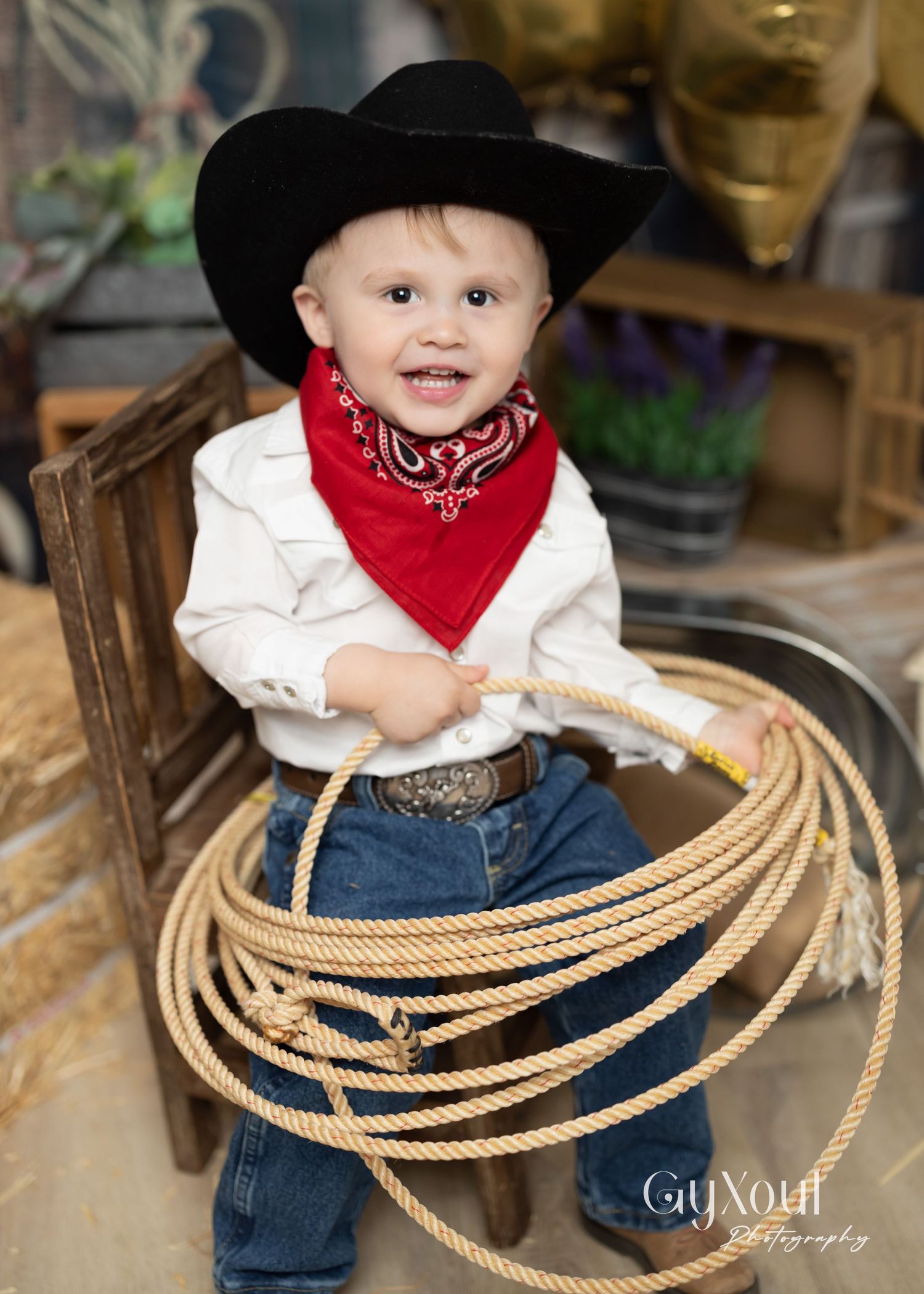 Young child in cowboy attire sits surrounded by western-style lasso ropes in an indoor photo session setup.