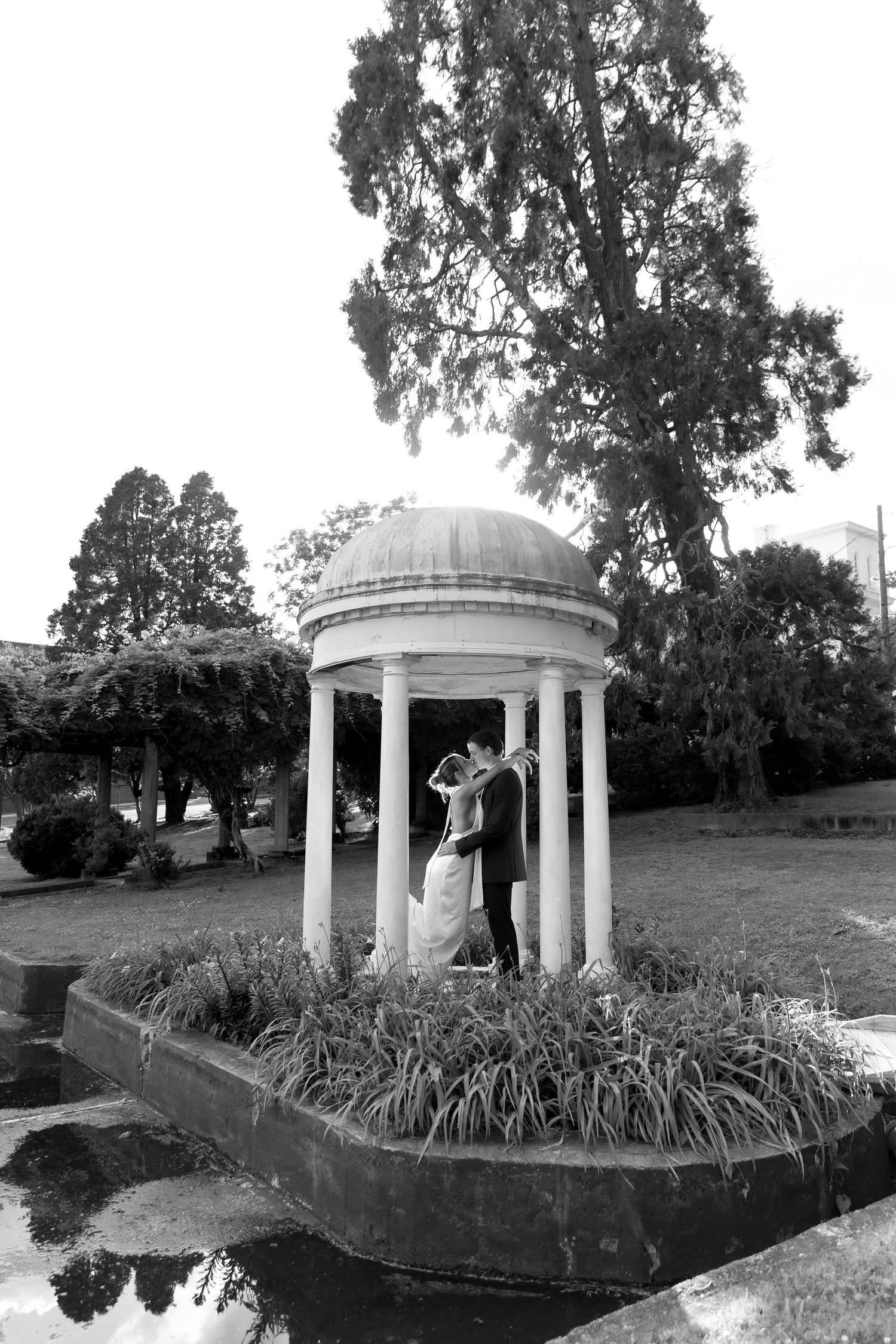 Black and white wedding photo of bride and groom embracing under a white domed gazebo surrounded by lush gardens.