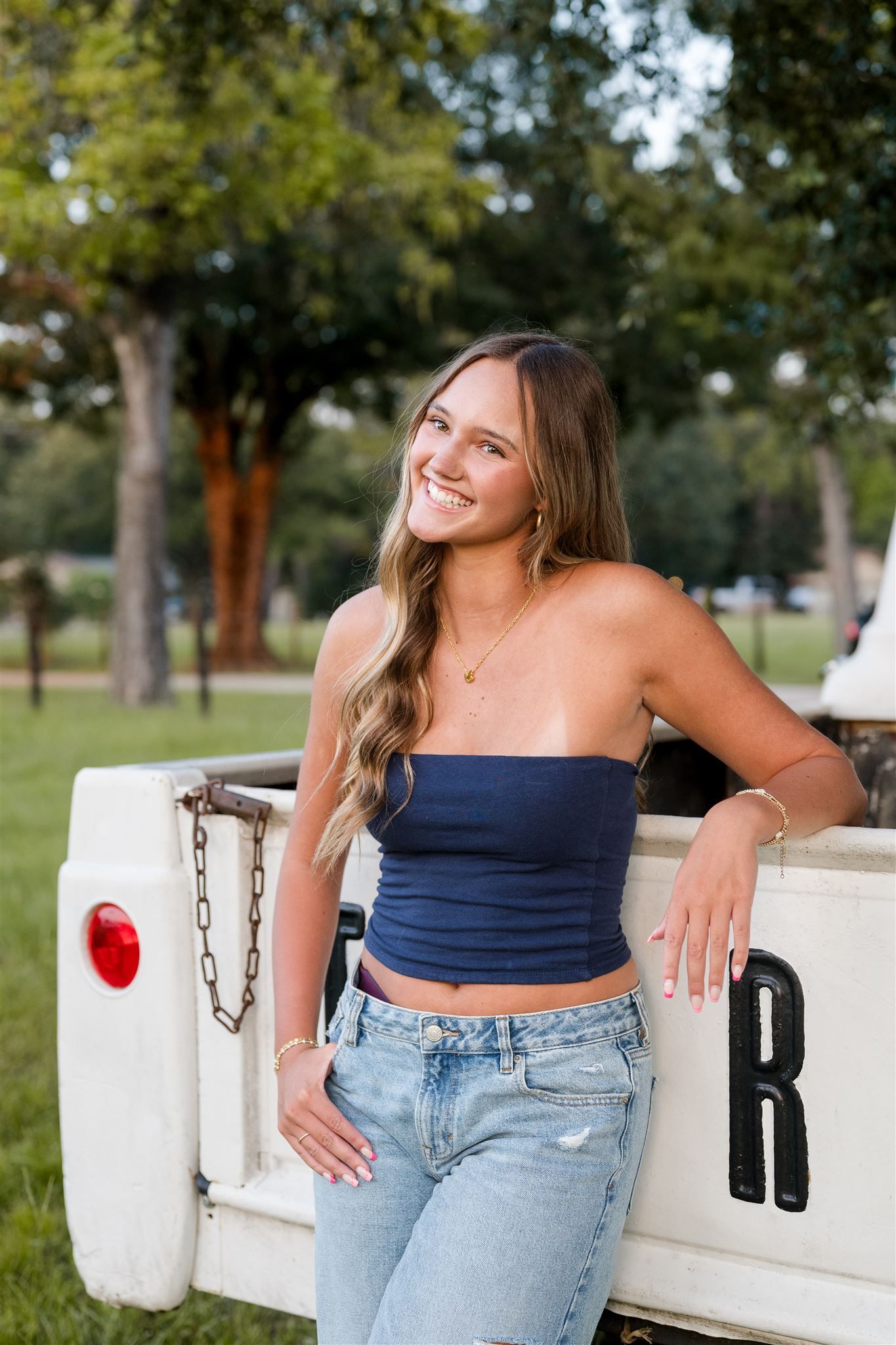 High school senior girl in navy blue tube top and light wash jeans poses casually against a white truck in an outdoor setting.
