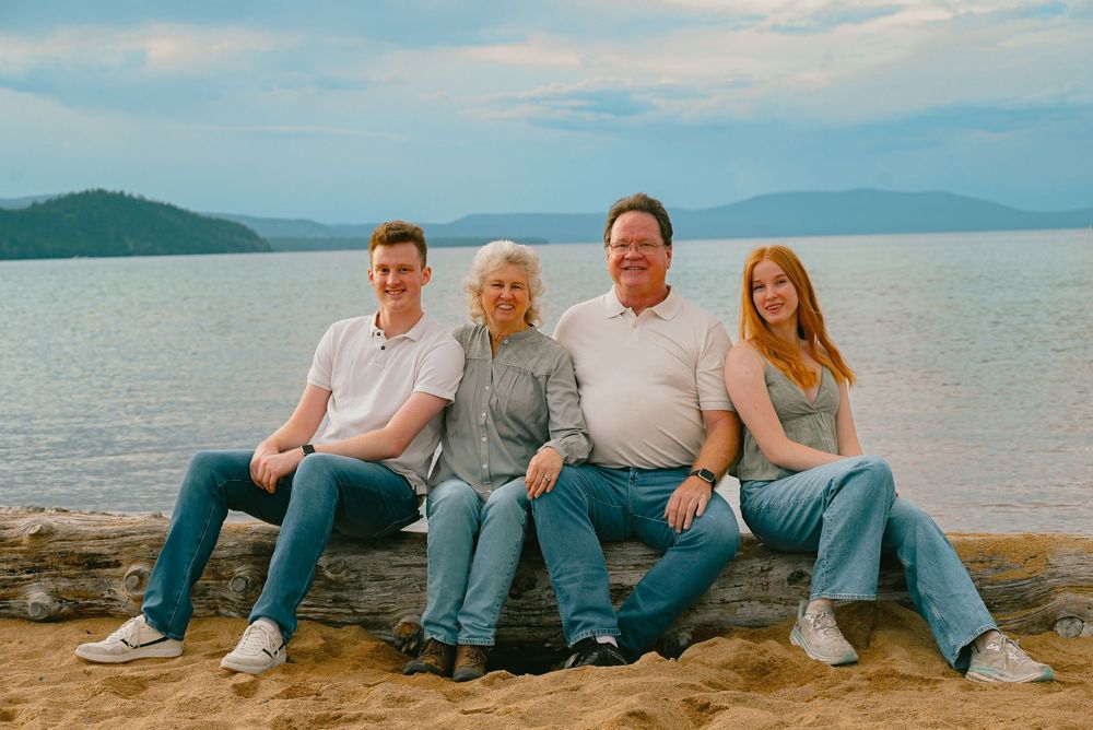 A group sits together on a beach with a scenic mountain lake view in the background on a sunny afternoon.