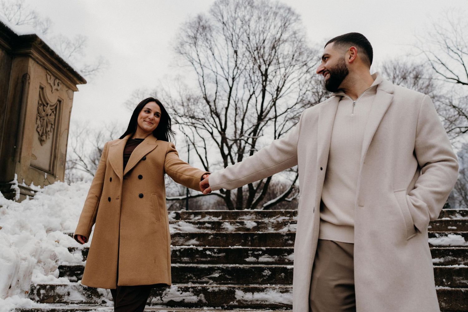 A couple playfully interacting on snow-covered stone steps in winter coats in Central Park