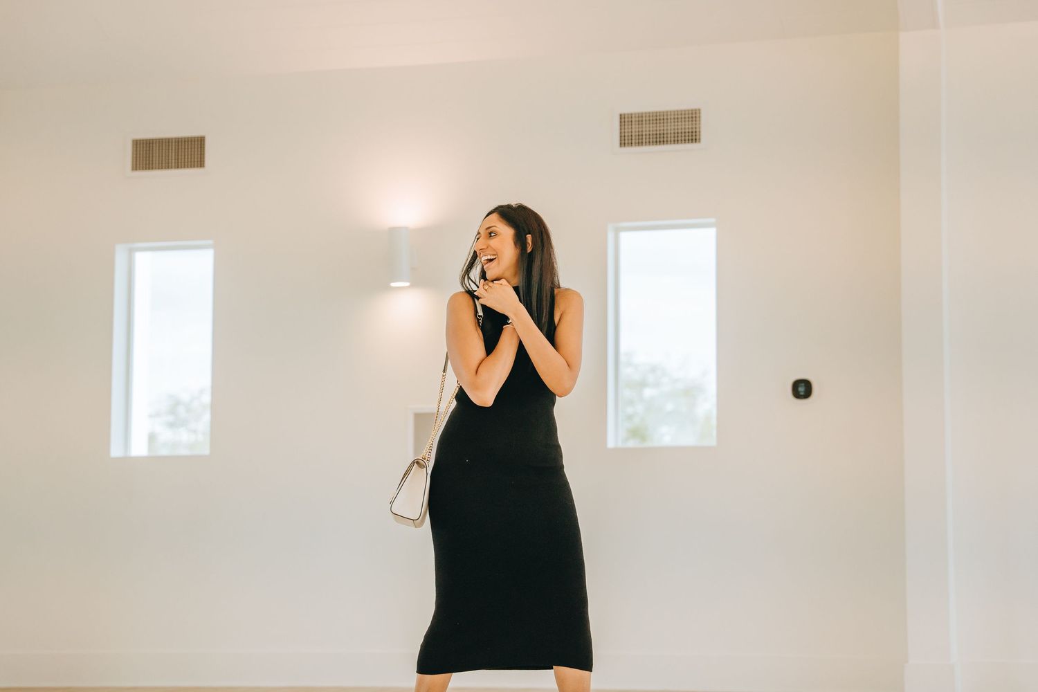 Woman in black dress stands in minimalist white interior with window lighting.