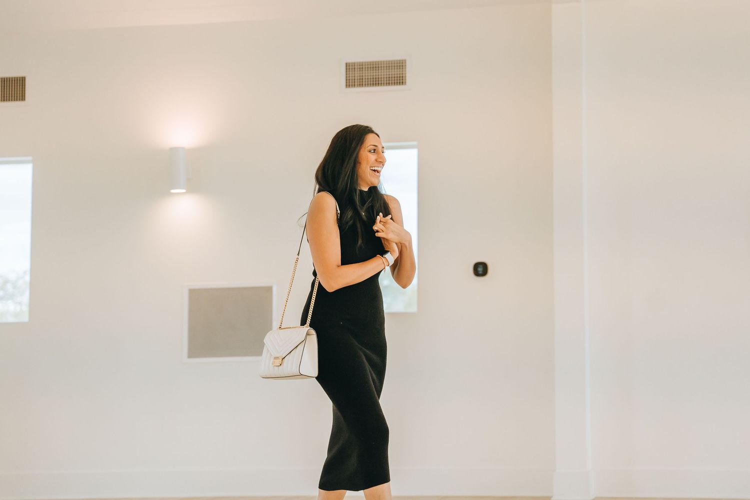 Woman in elegant black dress poses in bright white room with natural lighting.