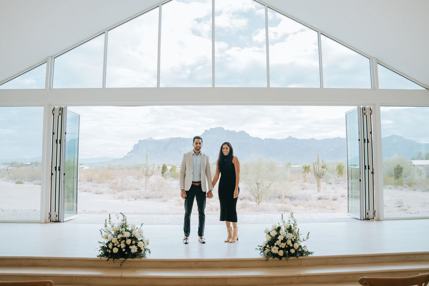 Couple standing on platform in modern white chapel with desert mountain backdrop.