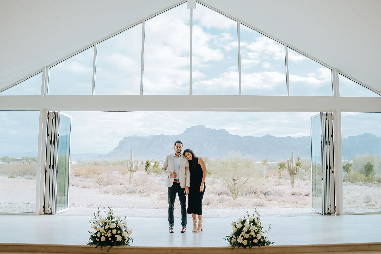 Couple standing in modern white chapel with desert landscape and floral decorations.