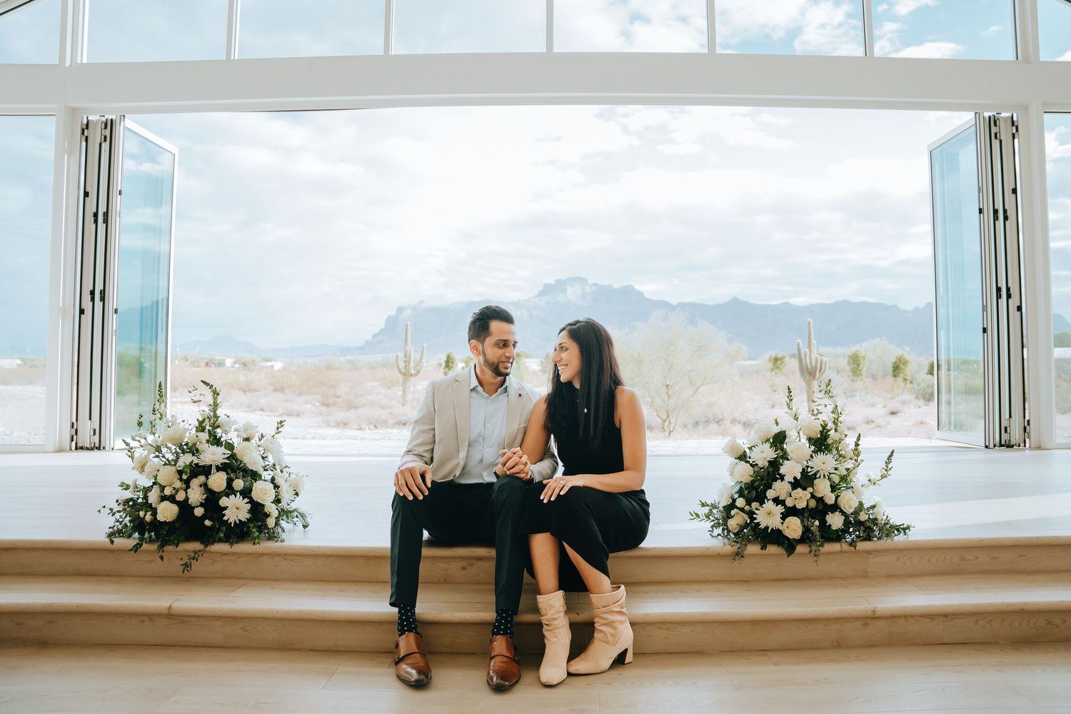 Couple sitting casually in modern chapel with desert mountain backdrop and white flowers.