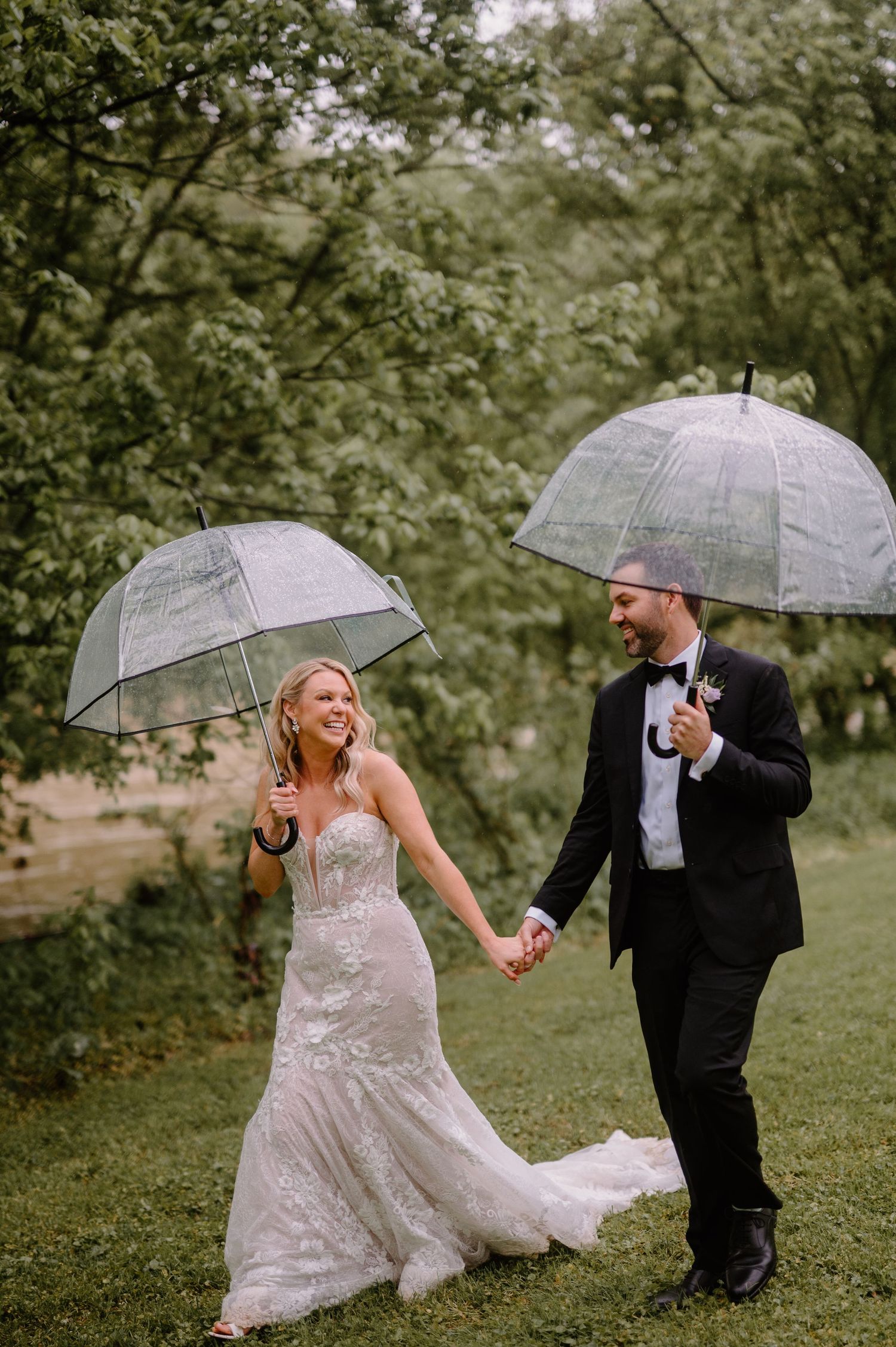 A bride and groom walk together holding hands while carrying clear umbrellas on a rainy wedding day in a garden setting.