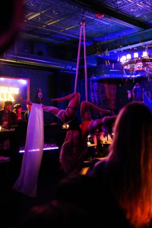 An aerial performer in white fabric performs gracefully against a blue-lit nightclub backdrop with chandeliers.