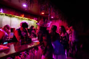 Crowded nightclub bar scene illuminated in vibrant pink and purple lighting.