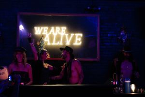'We Are Alive' neon sign glows above patrons in a dark nightclub atmosphere.