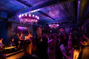 Dark nightclub interior with circular chandeliers and crowd gathered near stage under moody lighting.