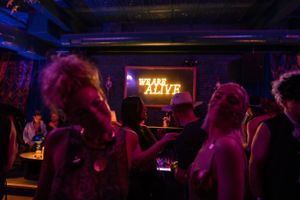 Silhouettes of clubgoers beneath neon 'ALIVE' sign in a dimly lit venue.