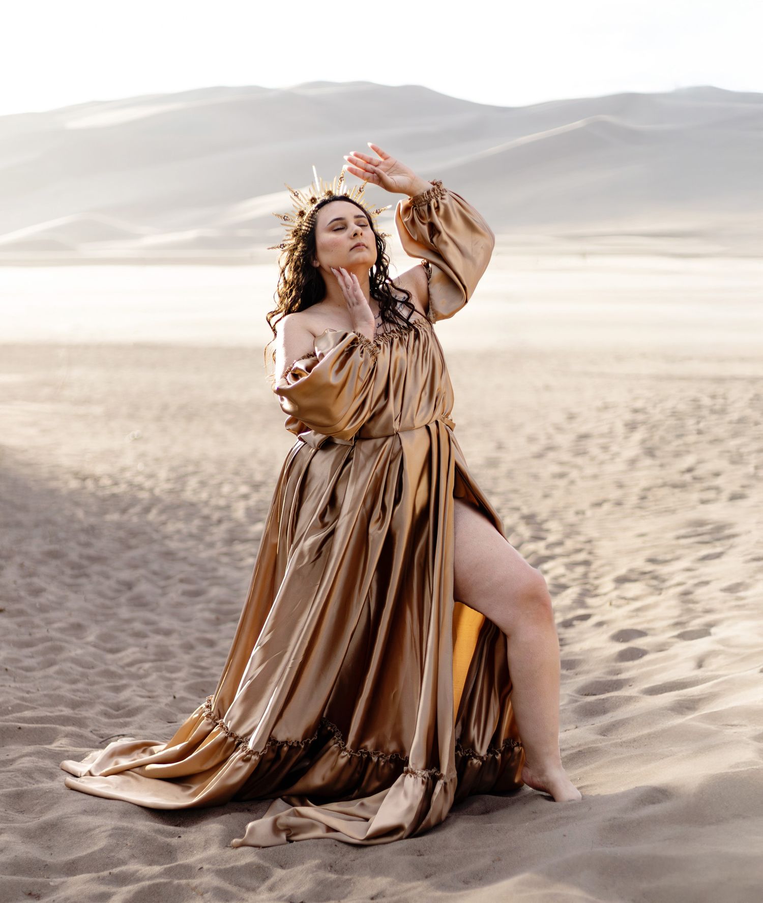 Woman in flowing tan dress poses gracefully on sandy beach during golden hour photography session.