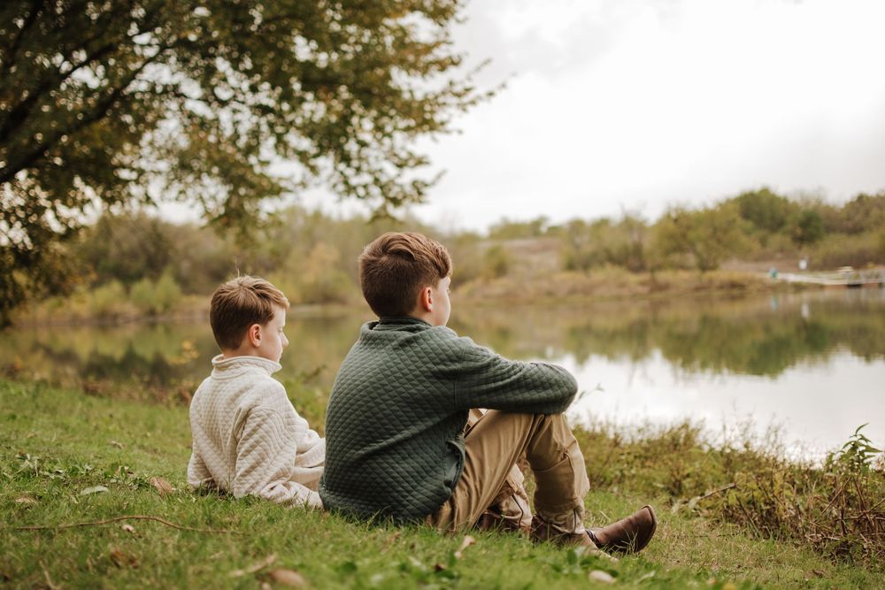 Two figures sit together peacefully on grassy hill overlooking a tranquil lake on an autumn day. Portrait Photographer Texas