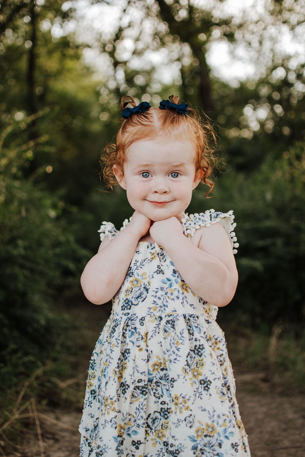 Child in floral dress poses sweetly outdoors with hands on cheeks.