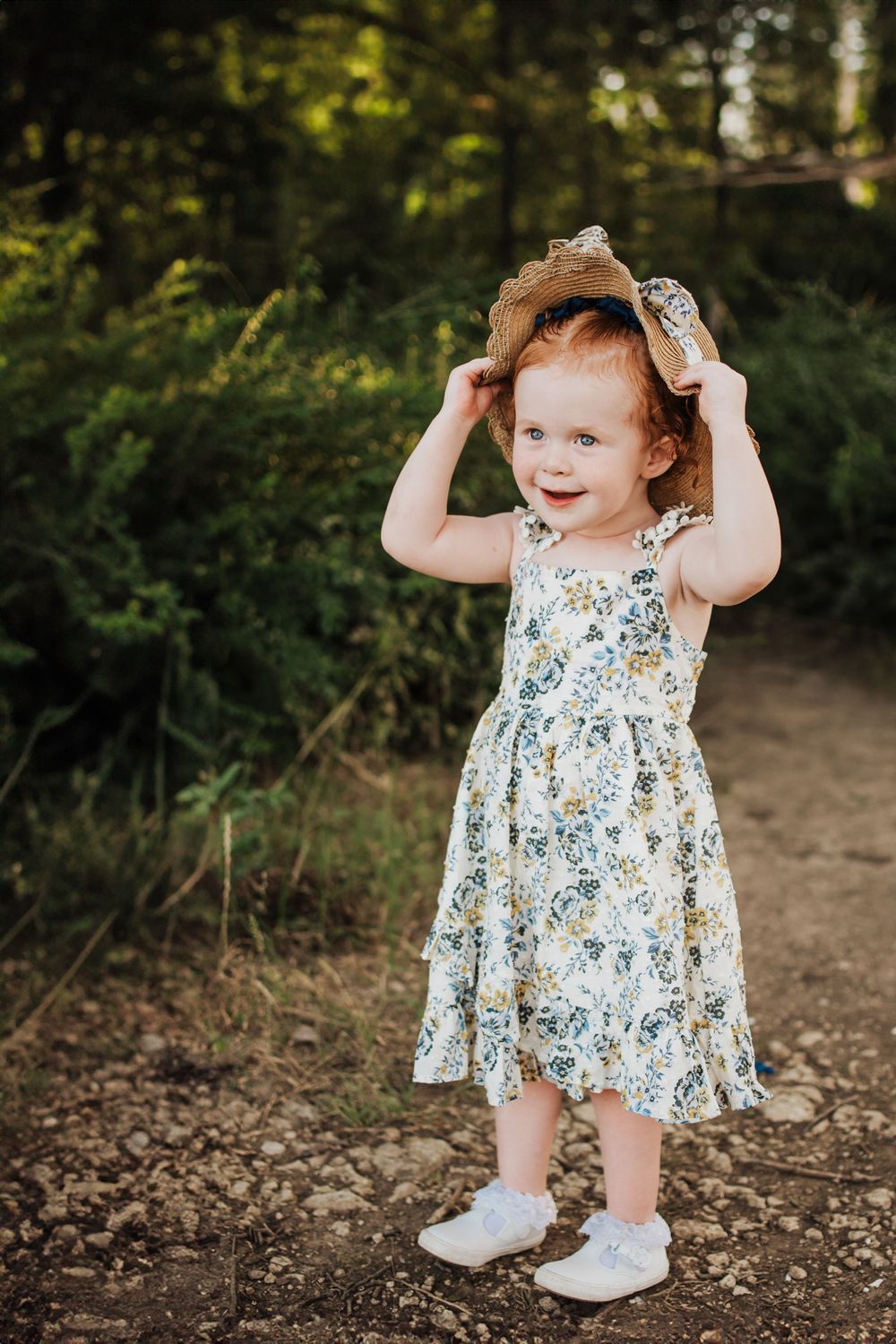 Toddler in floral dress plays with hair while standing on dirt path.