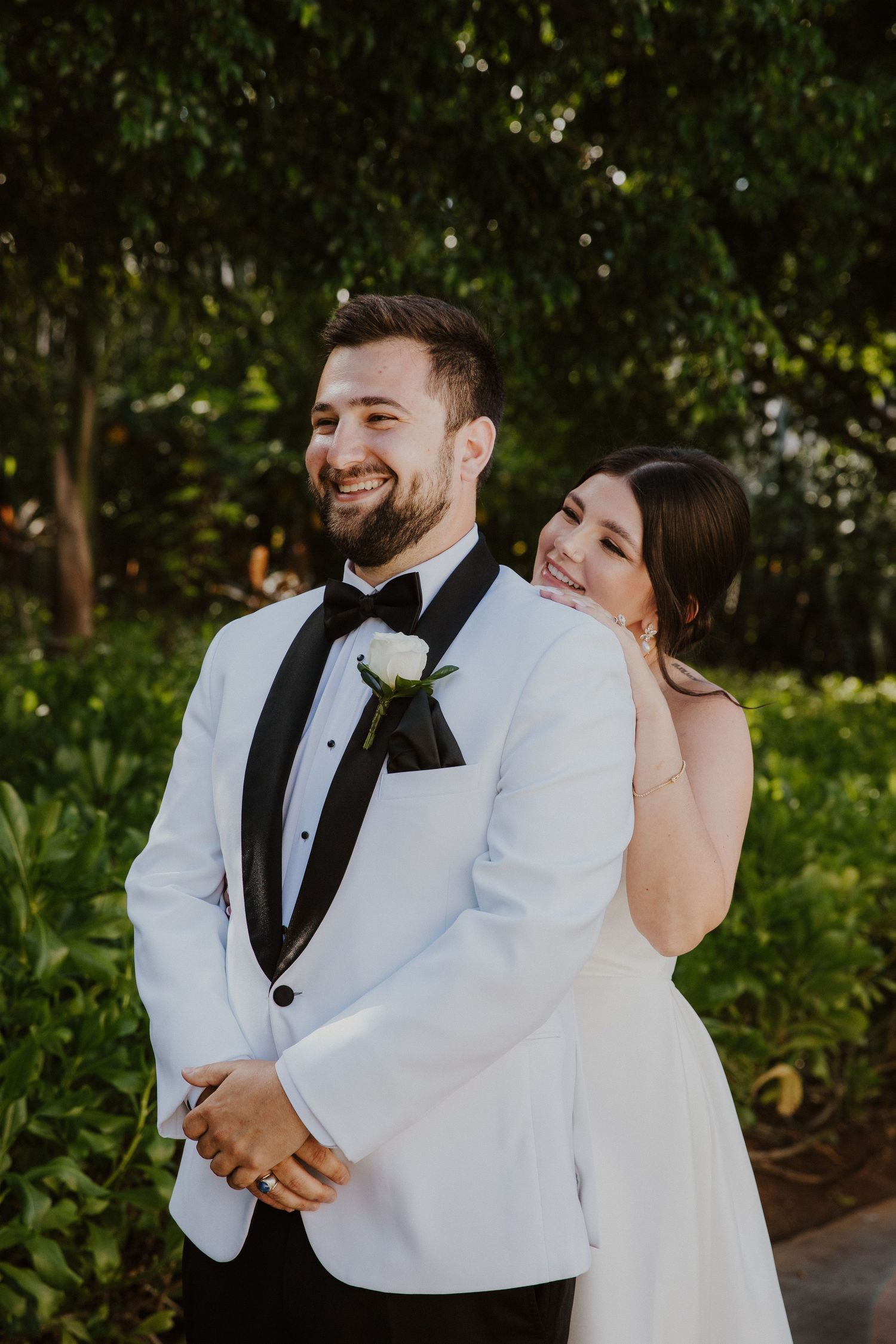 A bride and groom share a joyful moment together outdoors, with the groom wearing a white tuxedo jacket with black trim.